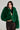 A woman in the vibrant green LUCKY HOODED FUR BOMBER and a black, semi-transparent polka-dot dress poses against a plain background. She has long dark hair and is looking slightly to the side.