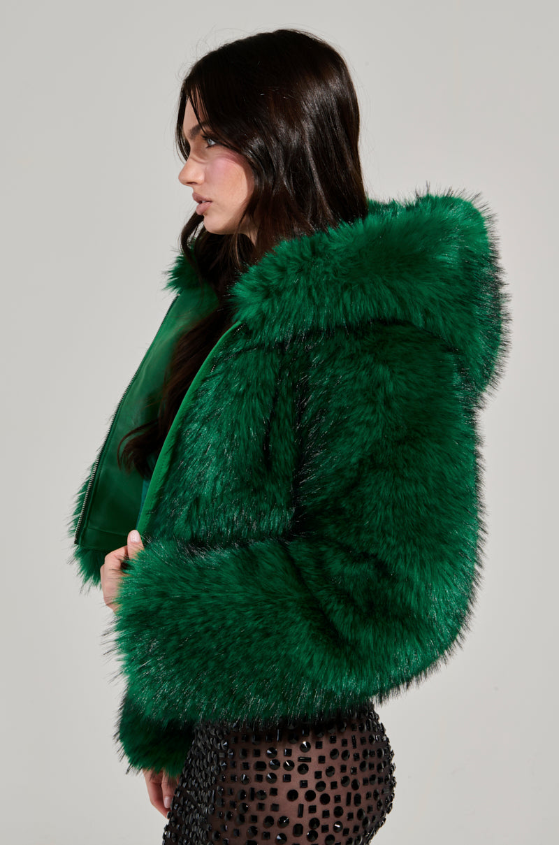 A woman with long dark hair stands in profile, wearing the eye-catching LUCKY HOODED FUR BOMBER in a vibrant green. She elegantly pairs it with a black skirt adorned with beads against a plain, light gray background.