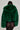 Wearing the LUCKY HOODED FUR BOMBER, she showcases her back in a green faux fur jacket, perfectly paired with a sheer black skirt embellished with circular designs, all elegantly framed by a neutral backdrop that enhances her look.