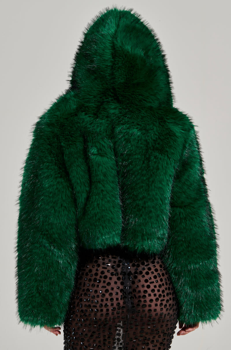 A person wearing the LUCKY HOODED FUR BOMBER, featuring a large and fluffy green hood, faces away from the camera. The jacket stylishly covers the upper body, while a sheer black skirt with circular designs is revealed below.