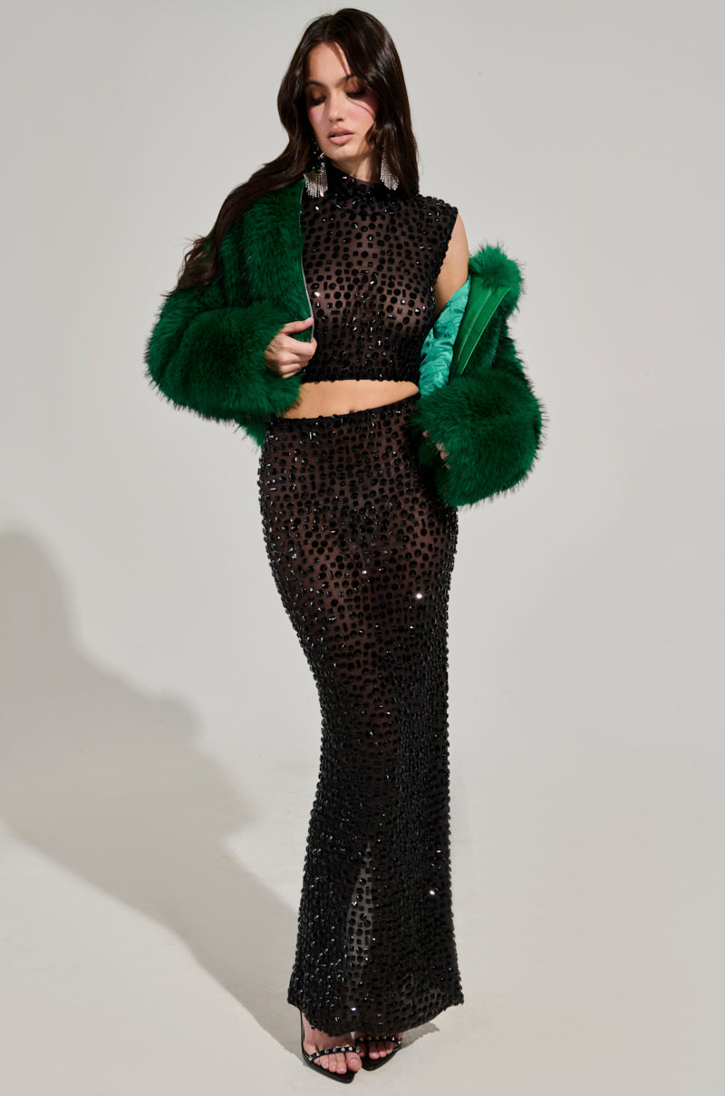 A woman poses in a stylish outfit featuring a sheer, black beaded dress with a high neckline, complemented by the LUCKY HOODED FUR BOMBER. She completes the look with black strappy high heels.