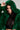 A person wearing the LUCKY HOODED FUR BOMBER in vibrant green with a black sequined top underneath gazes directly at the camera, holding the hood with one hand. The neutral background accentuates the stunning outfit.