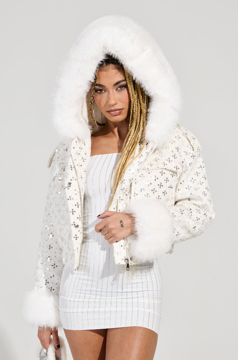 A person with long braids wears the SNOWBIRD FUR TRIM PUFFER jacket, a white hooded coat adorned with faux fur trim and sequin embellishments, over a white strapless dress while holding a small white purse.