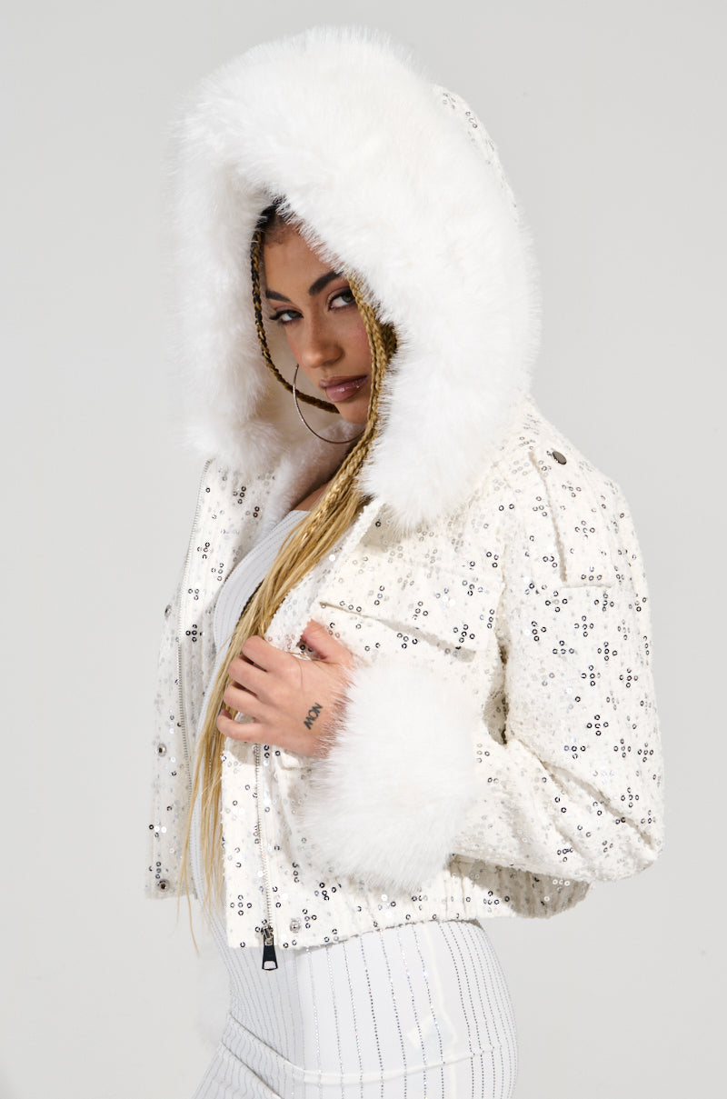 Wearing the SNOWBIRD FUR TRIM PUFFER, adorned with sequined embellishments, they have paired it with a stylish white outfit. The faux Sherpa lined interior provides warmth, and their long braided hair beautifully complements the chic ensemble set against a plain background.