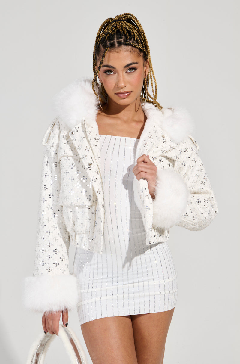 A woman with braided hair dons the sparkling SNOWBIRD FUR TRIM PUFFER, adorned with sequin embellishments, layered over a white, fitted dress. She carries a matching white handbag while the plain background highlights her fashionable ensemble and confident pose.