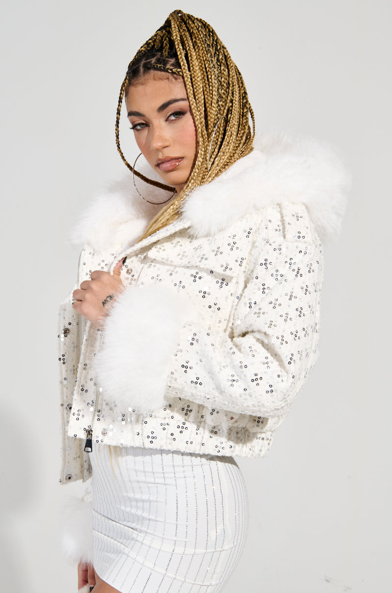 A person with braided hair confidently poses in the SNOWBIRD FUR TRIM PUFFER, its faux sherpa-lined interior adding warmth. Sequined embellishments shimmer on the white, fur-collared jacket as they stand against a plain background, wearing a white striped skirt and hoop earrings.