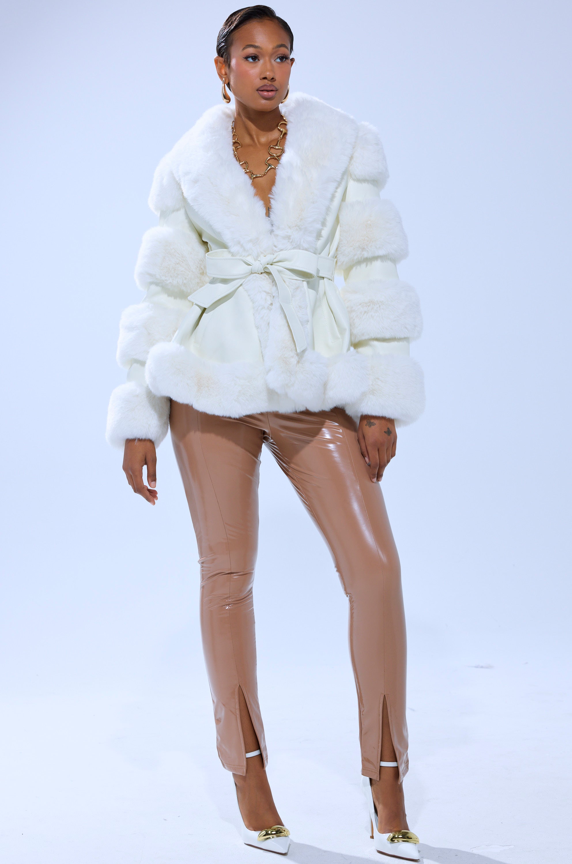 A woman stands confidently in the WINTER WONDERLAND FUR TRIM MOTO, shiny beige pants with ankle front slits, white heels, and gold jewelry against a plain light background.