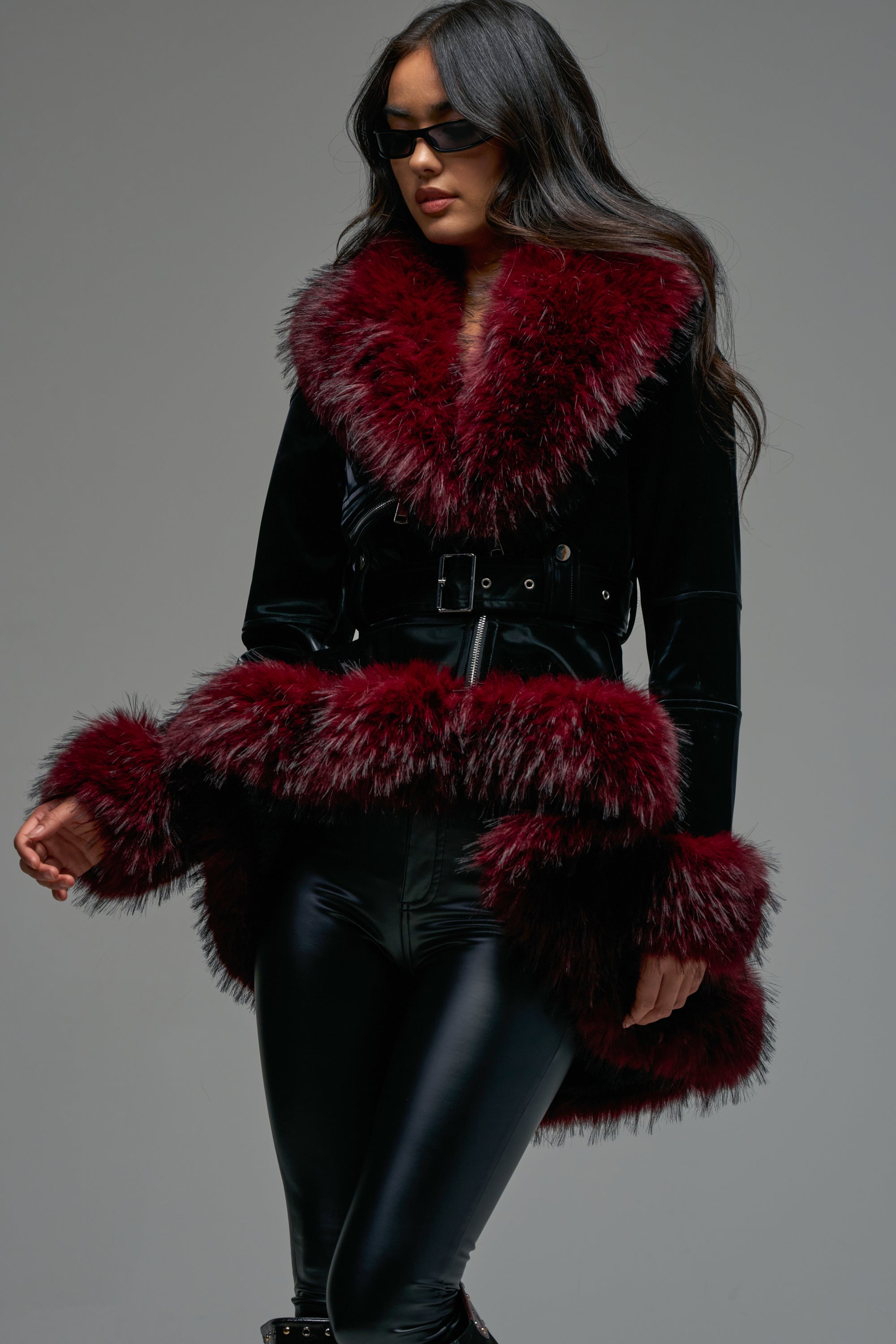 A woman wearing the DO NO WRONG FUR TRIM MOTO, a belted velvet coat with large burgundy fur trim on the collar, cuffs, and hem, styled over shiny black leggings. She has long hair and sports black sunglasses.
