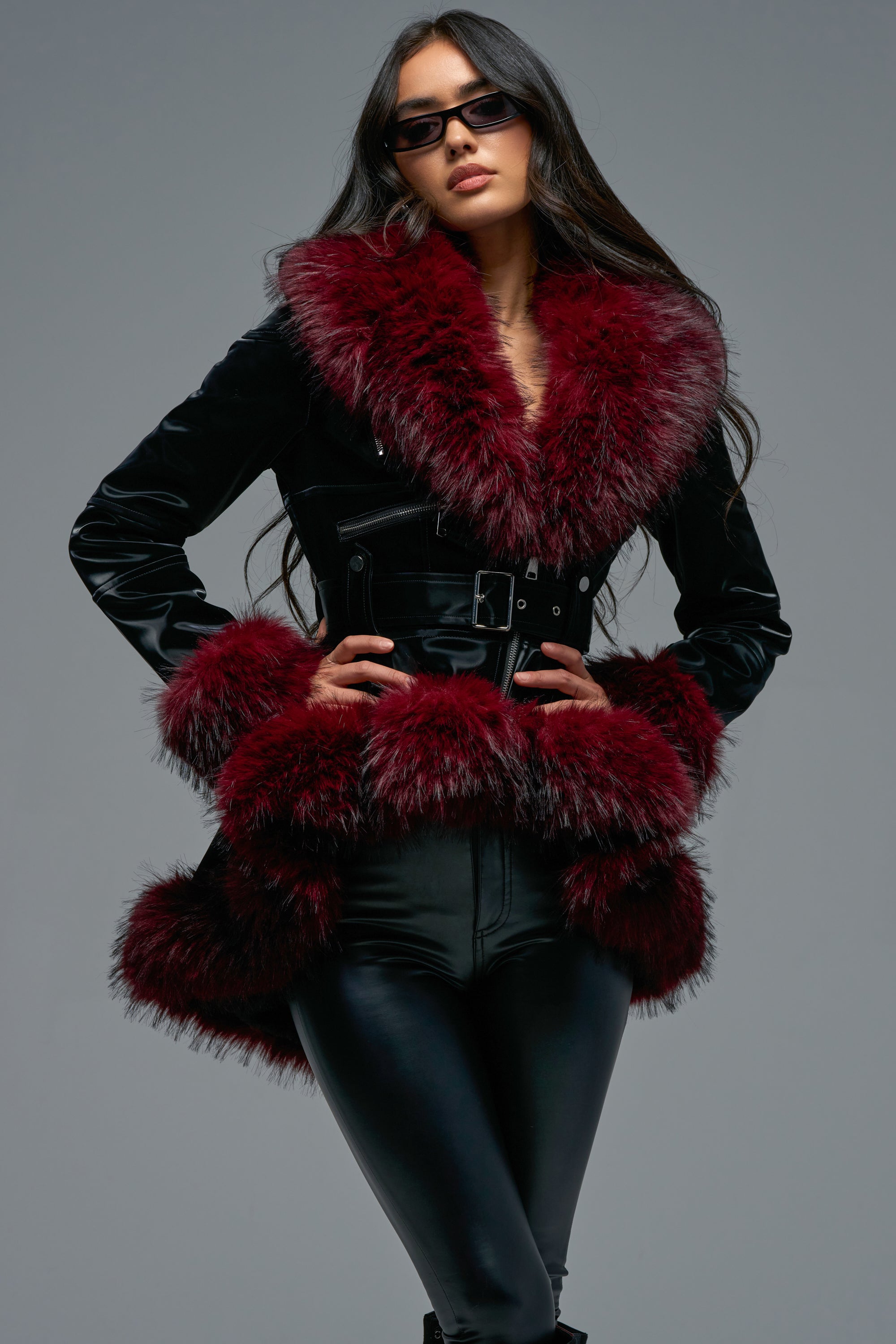 Wearing the DO NO WRONG FUR TRIM MOTO, a woman in black sunglasses stands confidently against a plain gray background. The black moto coat features a striking belted design with large, dark red faux fur trim at the collar, cuffs, and hem. Paired with black leather pants for a bold ensemble, its peplum silhouette adds an elegant finishing touch to her chic look.