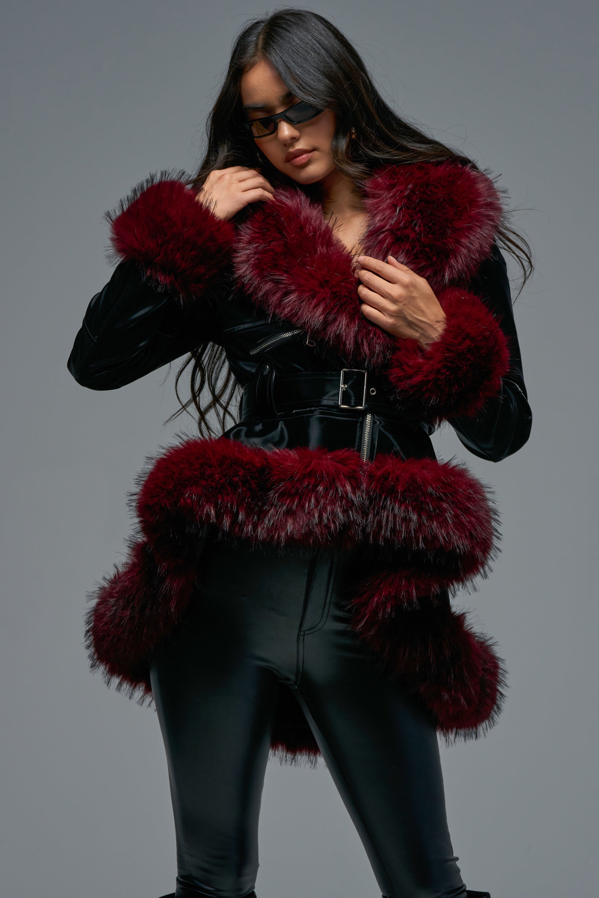 A woman poses against a gray background wearing black sunglasses, black pants, and the DO NO WRONG FUR TRIM MOTO—a black belted coat featuring a burgundy fur trim.