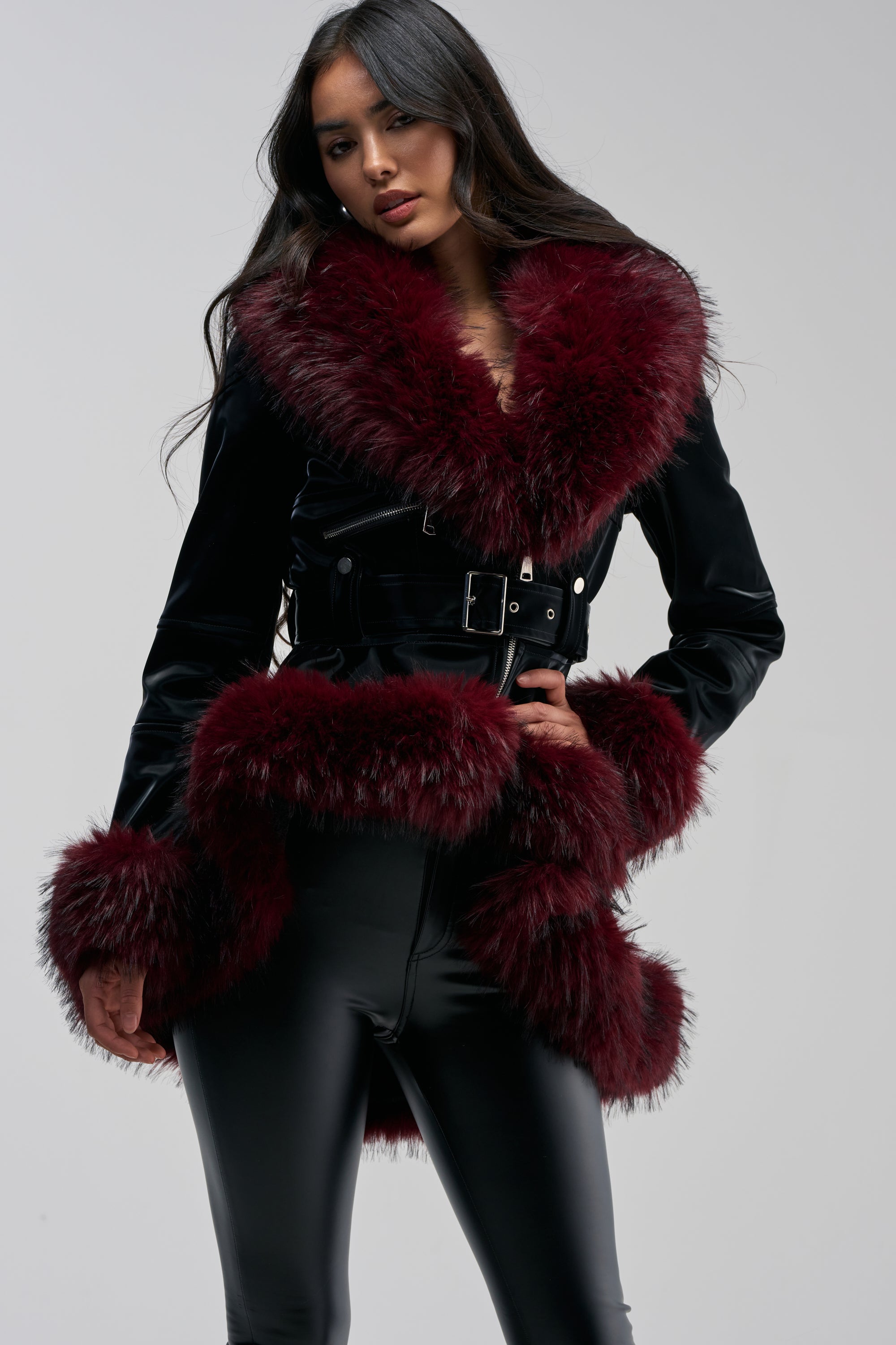 A woman models the DO NO WRONG FUR TRIM MOTO, a black coat adorned with oversized burgundy faux fur at the collar, cuffs, and hem, showcasing a chic peplum silhouette. She pairs it with shiny black pants and poses against a simple light background.