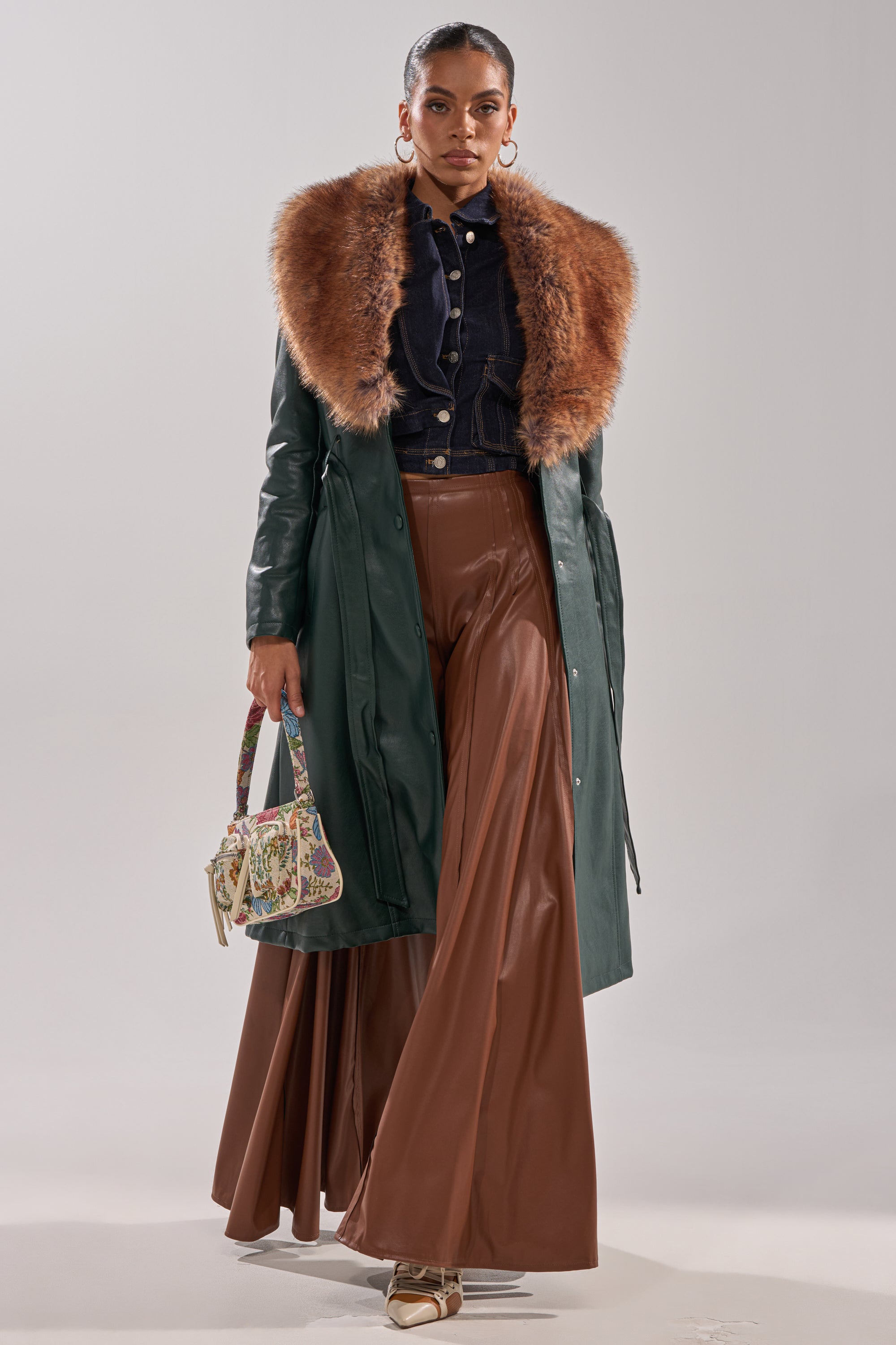 Wearing the GREENVIEW FUR TRIM TRENCH, a woman pairs it with wide-legged brown trousers, a black buttoned top, gold shoes, and a floral handbag as she stands confidently against a plain background.
