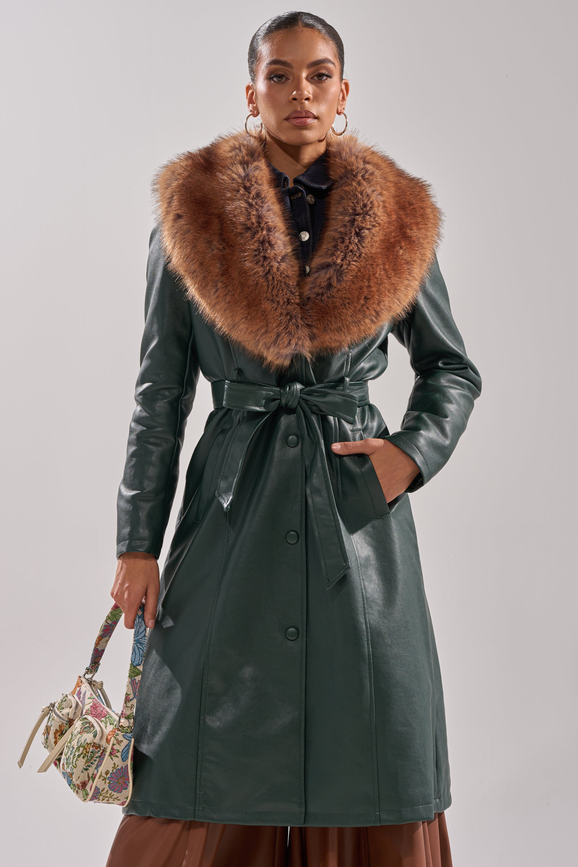 A woman stands facing forward in the GREENVIEW FUR TRIM TRENCH, a green coat featuring a wide brown fur-trimmed collar, holding a small patterned handbag with one hand in her pocket.