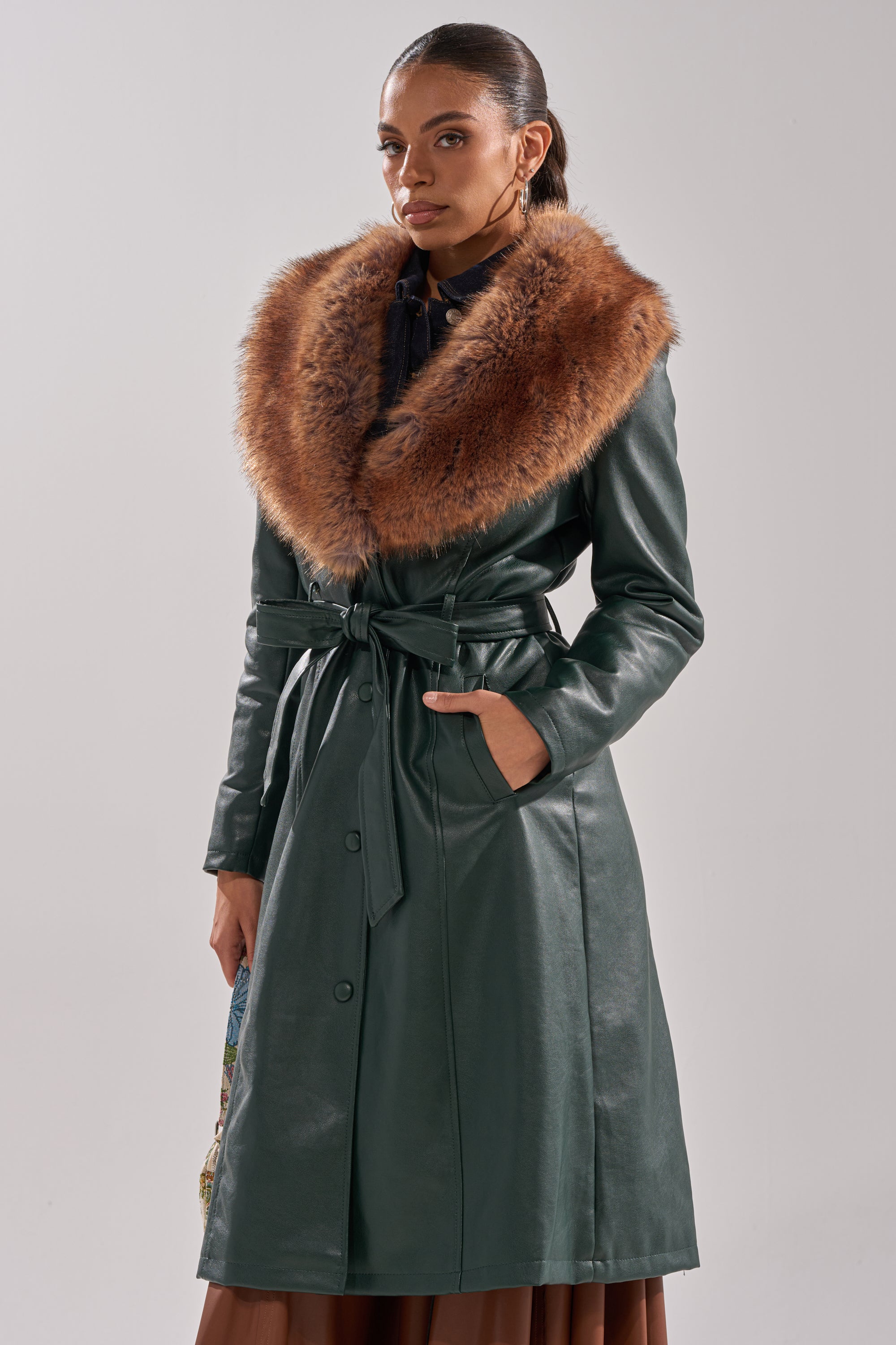 A woman models the GREENVIEW FUR TRIM TRENCH, a forest green faux leather coat with a wide brown fur collar and belted waist, posing with one hand in her pocket against a plain light background.