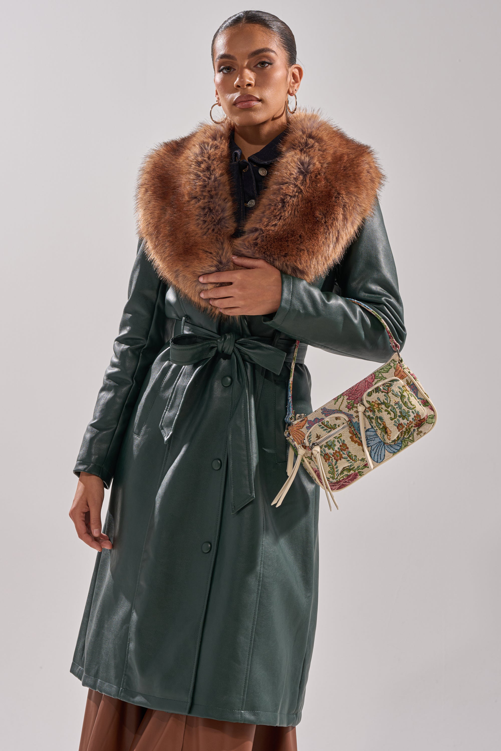 A woman in the GREENVIEW FUR TRIM TRENCH, featuring dark green faux leather with a large brown faux fur collar, hoop earrings, and a floral handbag, stands against a plain light background.
