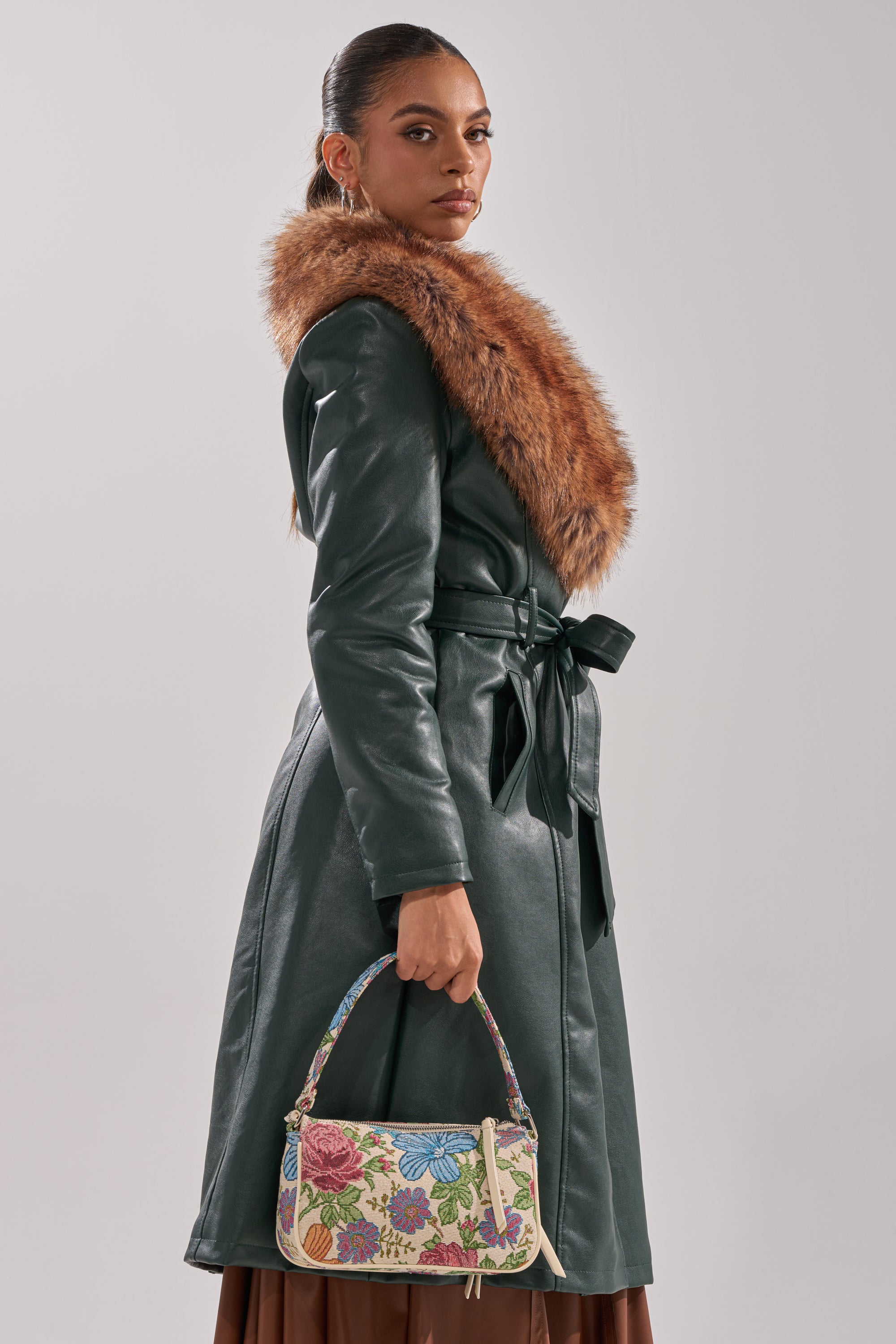 A woman wears the GREENVIEW FUR TRIM TRENCH, featuring a green coat with a brown fur collar. She holds a small floral handbag and stands sideways, gazing confidently at the camera against a plain light background.