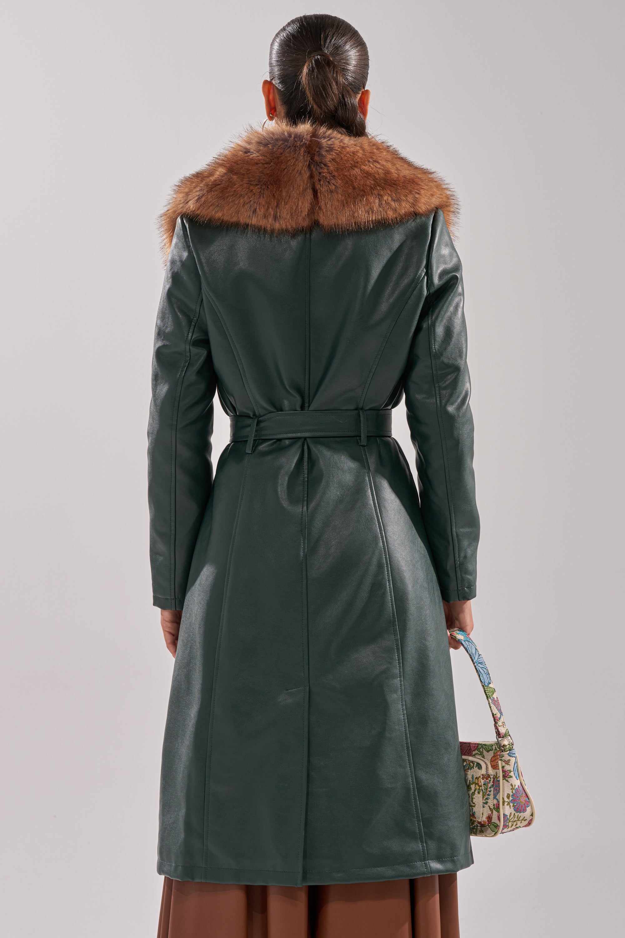 A woman with dark hair in a low ponytail stands with her back to the camera, wearing the GREENVIEW FUR TRIM TRENCH—a dark green faux leather coat with a brown fur collar—while holding a small patterned handbag.