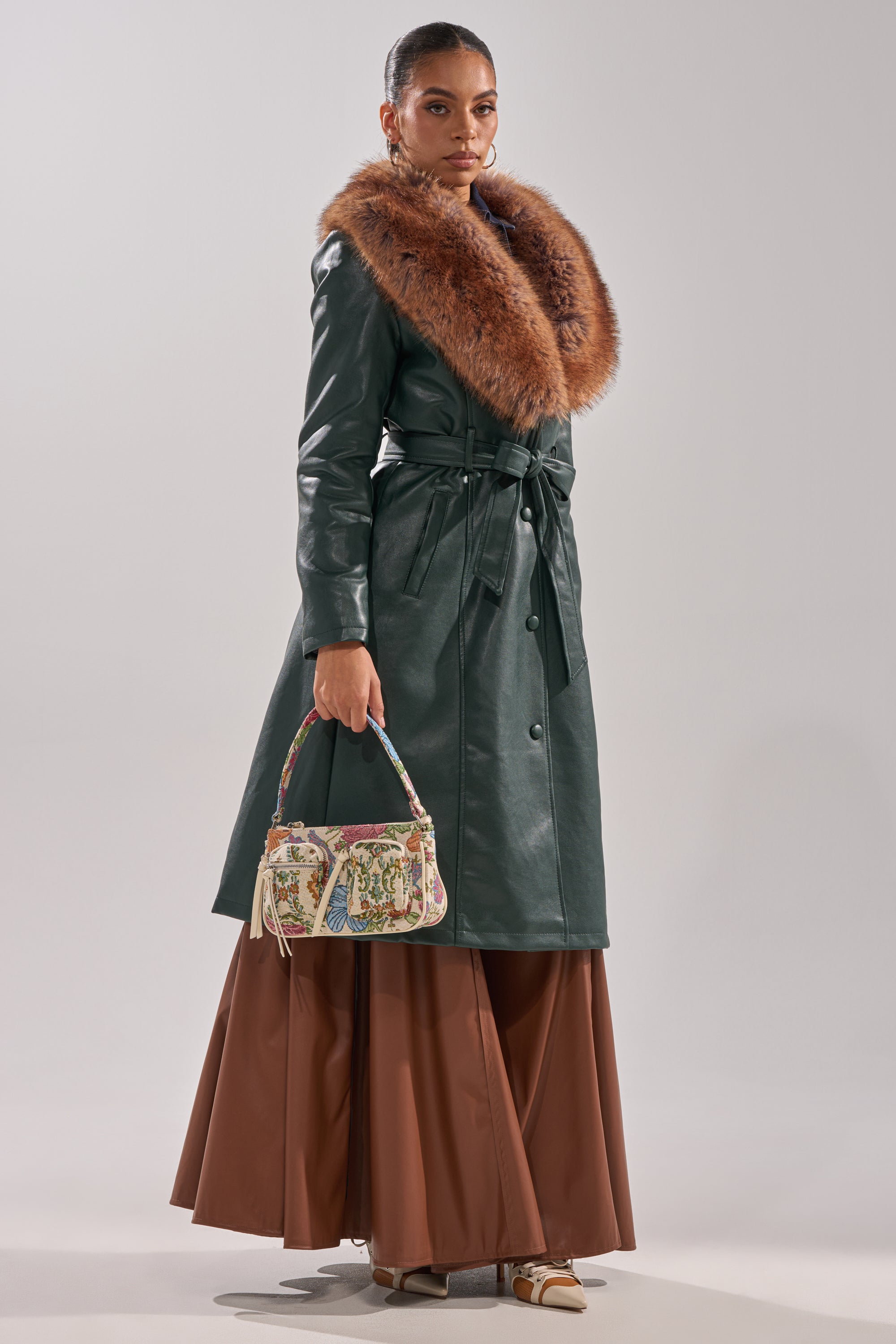 A woman models the GREENVIEW FUR TRIM TRENCH—a long green coat with a wide brown fur collar—paired with a brown maxi skirt, beige heels, and a small patterned handbag against a plain light background.