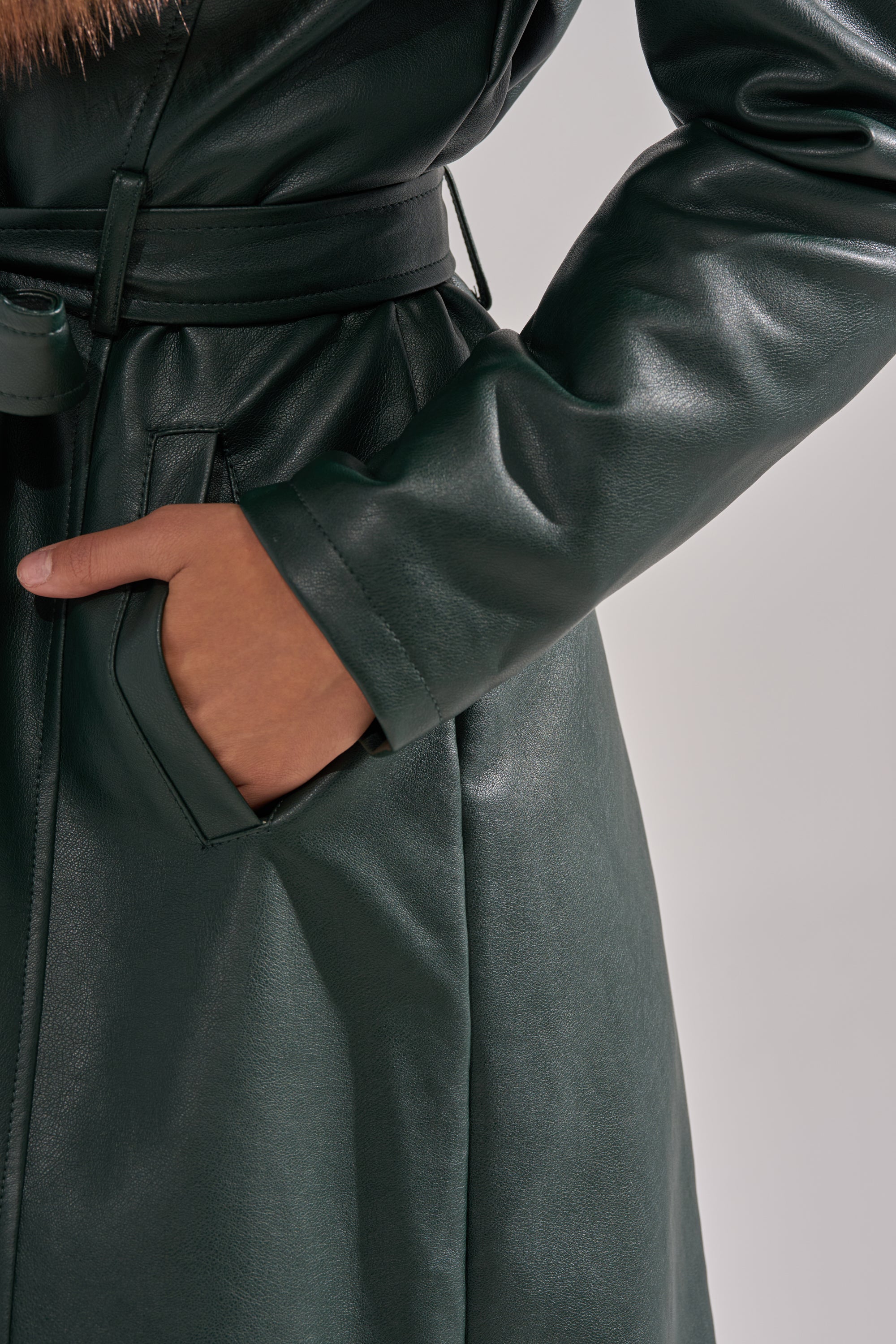 A person models the GREENVIEW FUR TRIM TRENCH—a dark green faux leather coat—shown from shoulders to mid-thigh, with one hand in the pocket against a plain background.