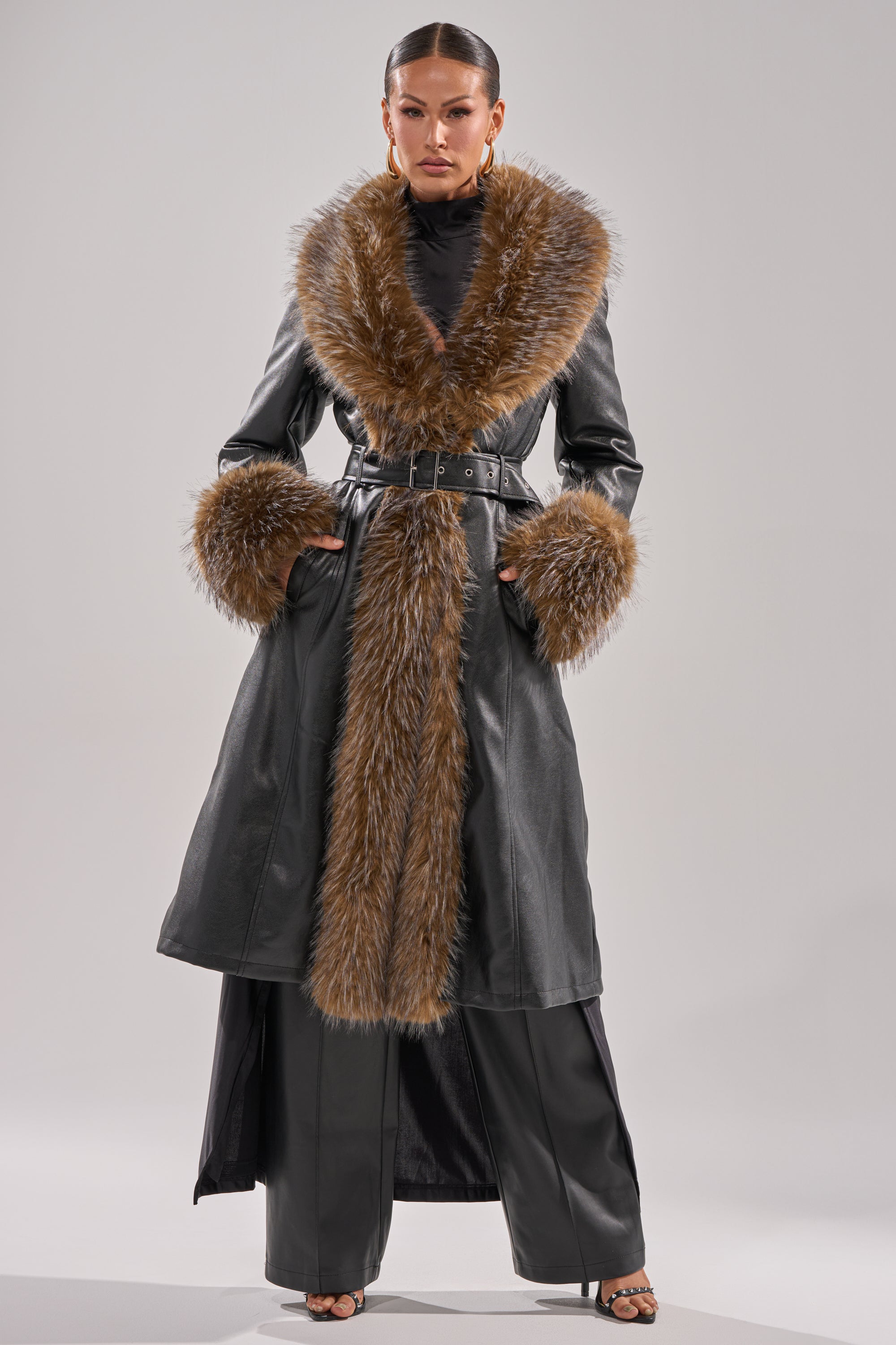 A woman faces forward in the VOODOO FUR TRIM TRENCH, featuring brown faux fur on the collar, cuffs, and front. It's belted at the waist and paired with black wide-leg pants and black heeled sandals.