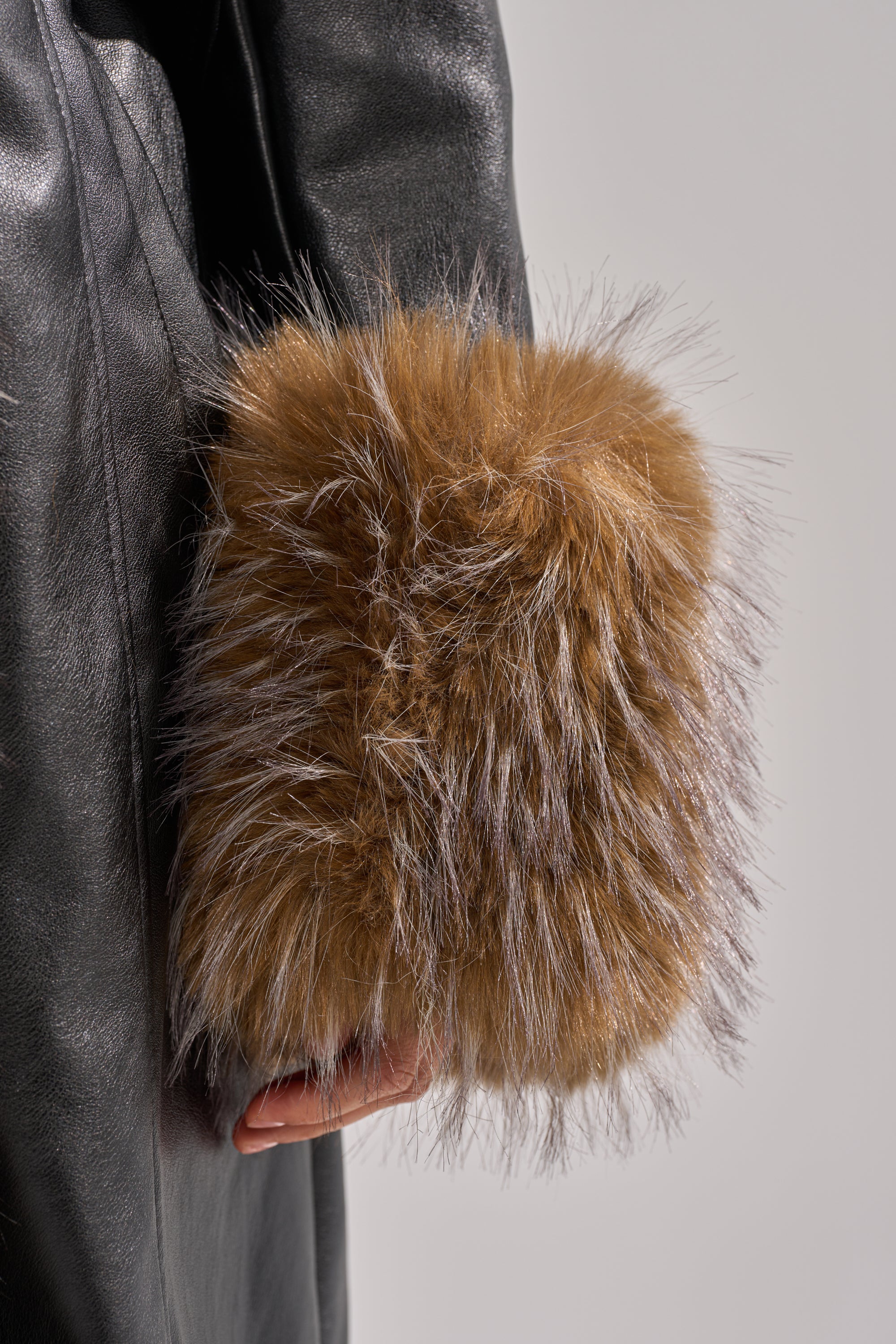 A close-up of a person wearing the VOODOO FUR TRIM TRENCH, a black faux leather coat with large brown faux fur trim cuffs. The longline silhouette highlights its luxurious texture as the person's hand peeks from under the cuff.