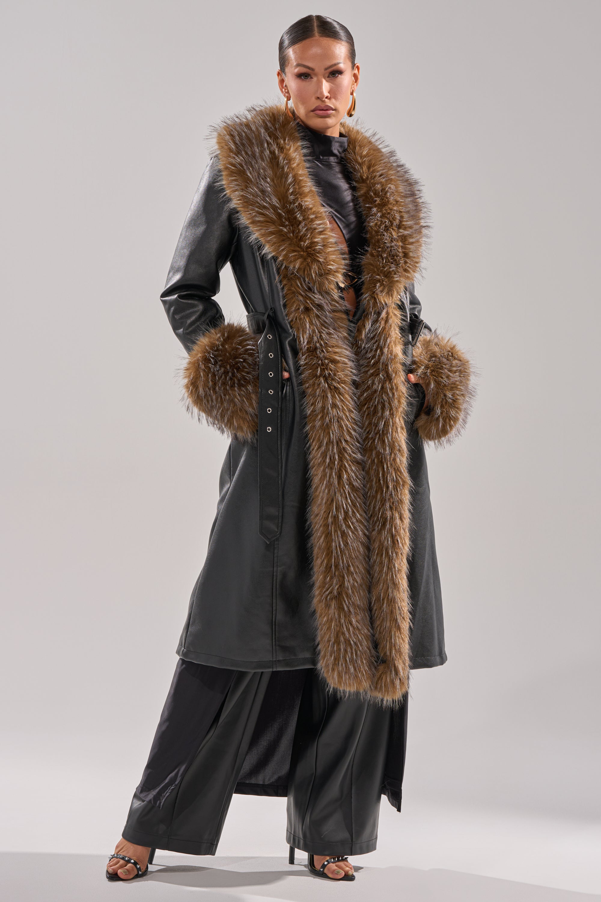 A person stands confidently in the VOODOO FUR TRIM TRENCH—a long black coat with wide brown faux fur collar and cuffs—paired with black pants and heeled sandals, set against a simple light background.