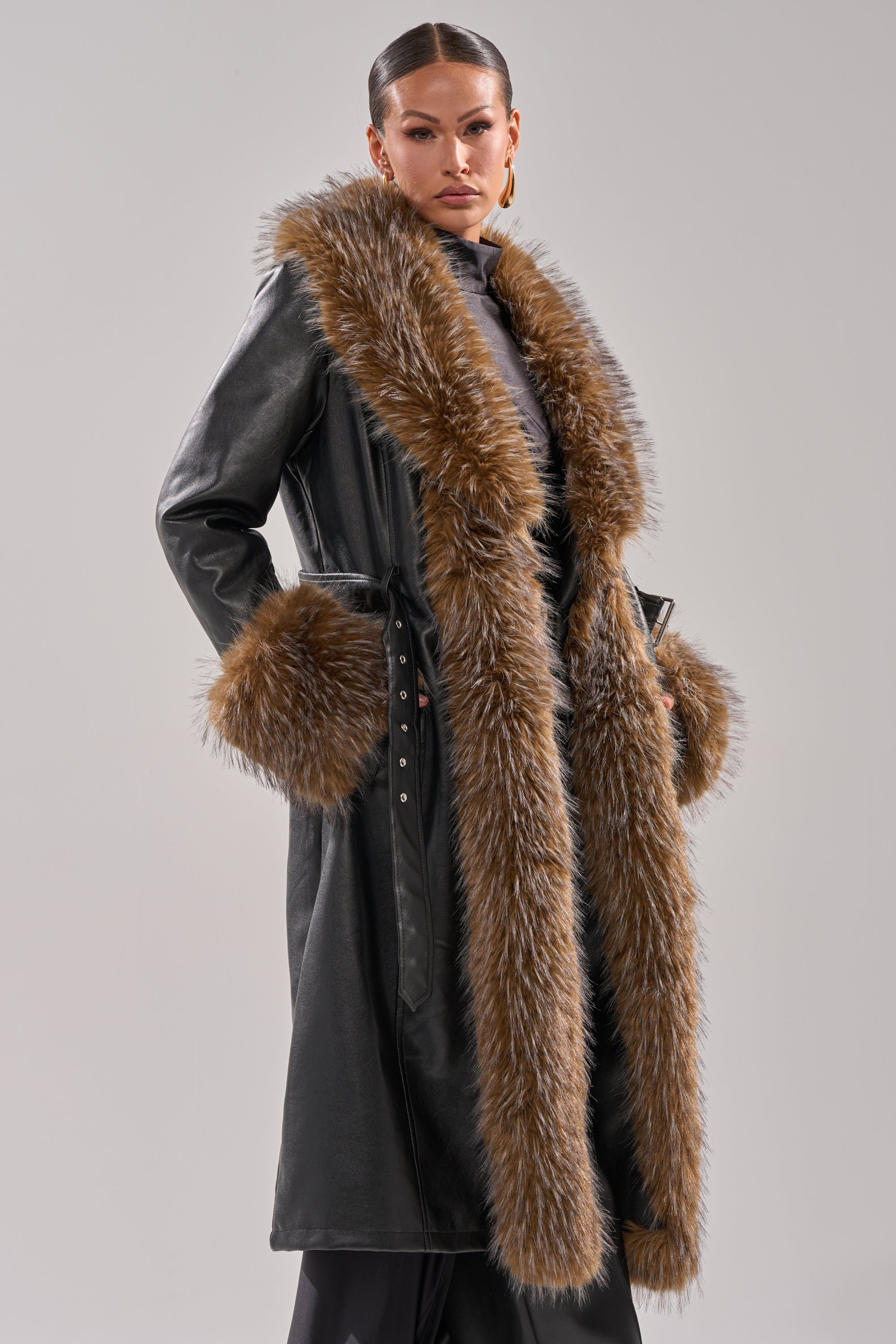 A woman stands confidently against a light gray background, wearing the VOODOO FUR TRIM TRENCH—a faux leather coat featuring brown fur trim on the collar, cuffs, and front edges.