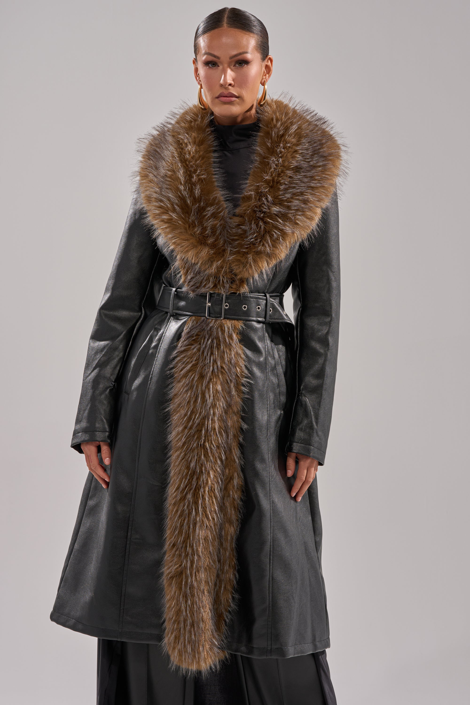 A woman models the VOODOO FUR TRIM TRENCH, a faux leather coat with a wide brown fur collar and belted waist, against a plain gray background. She has slicked-back hair and gold hoop earrings.