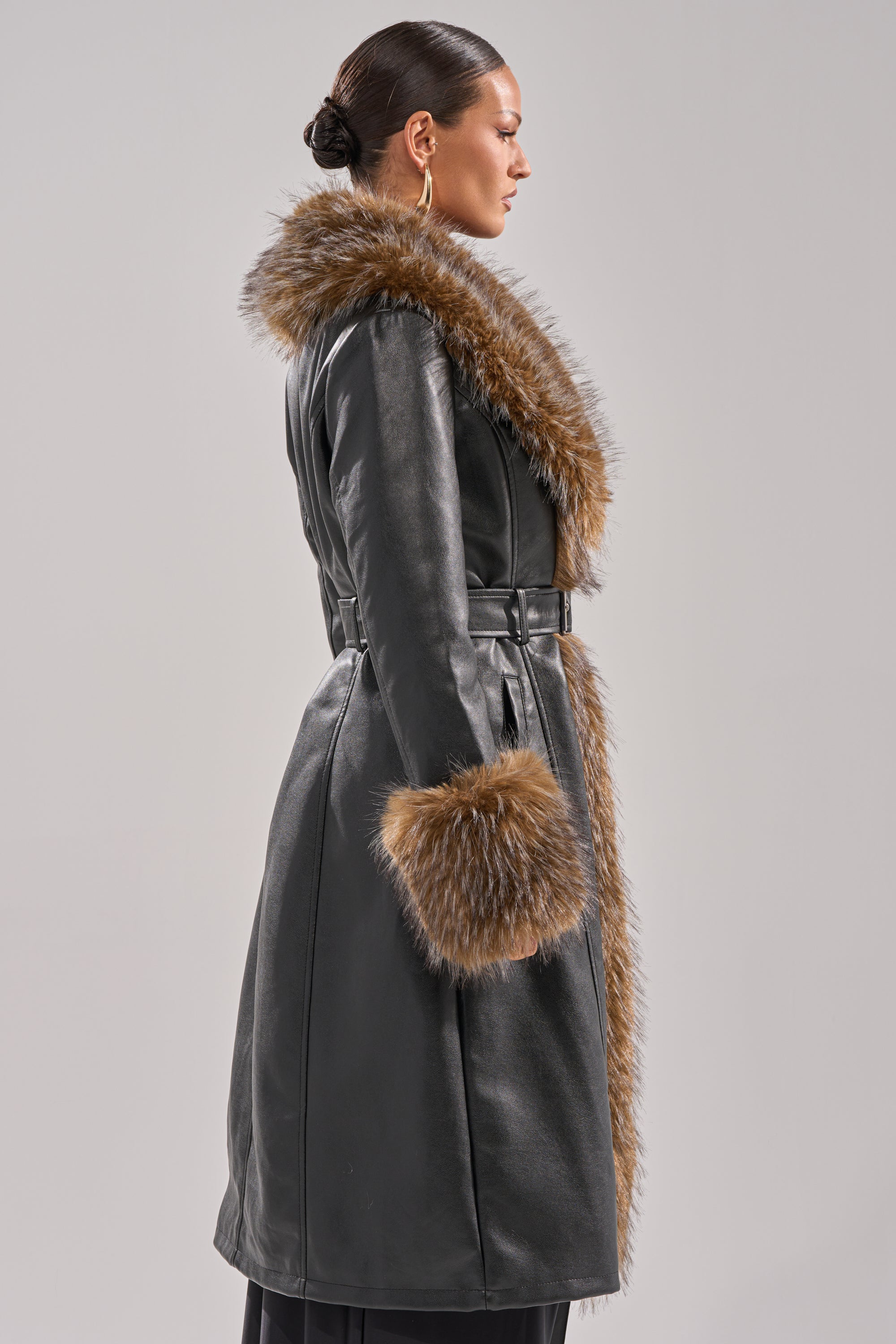 A woman stands in profile wearing the VOODOO FUR TRIM TRENCH, a faux leather coat with a wide brown fur collar and matching fur cuffs, her hair tied back and accessorized with large hoop earrings.