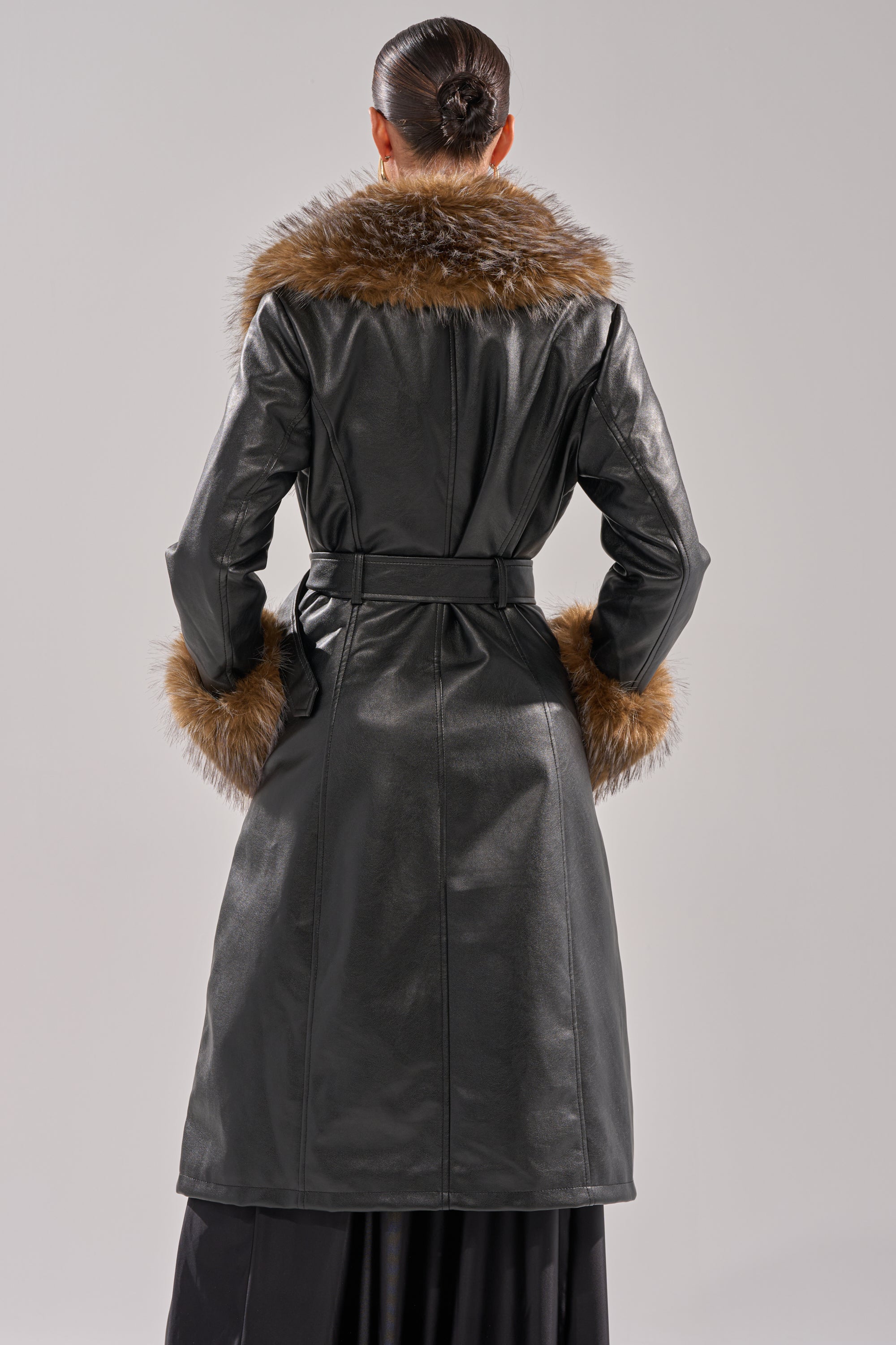 A person stands with their back to the camera, wearing the VOODOO FUR TRIM TRENCH—a long faux leather coat with brown fur on the collar and cuffs. Their hair is in a sleek bun against a plain, light-colored background.