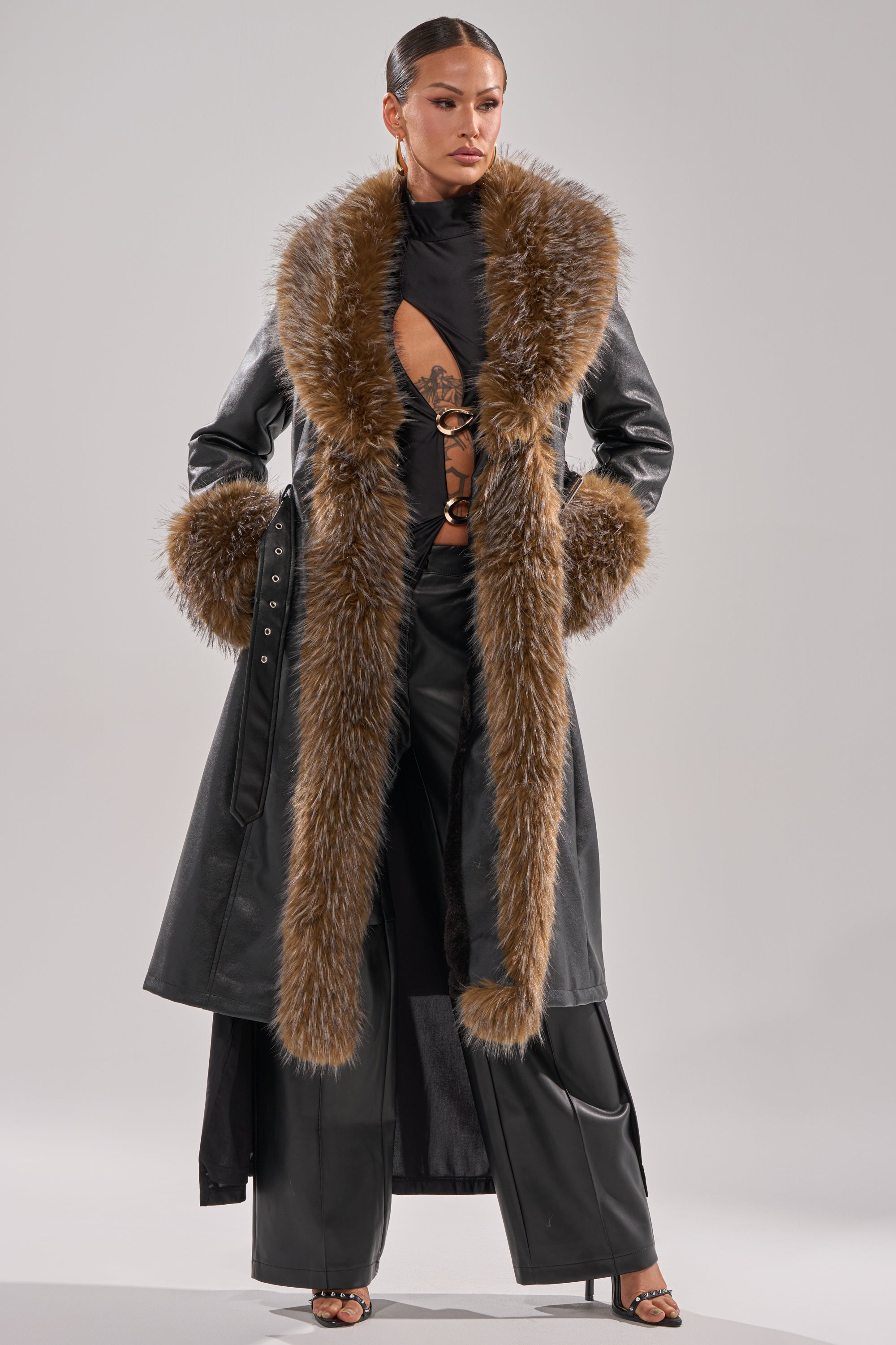 A woman exudes confidence in the VOODOO FUR TRIM TRENCH, a long faux leather coat with bold brown fur trim at the collar and cuffs, styled over a cut-out black top and wide-leg pants, her hair slicked back for a sleek look.