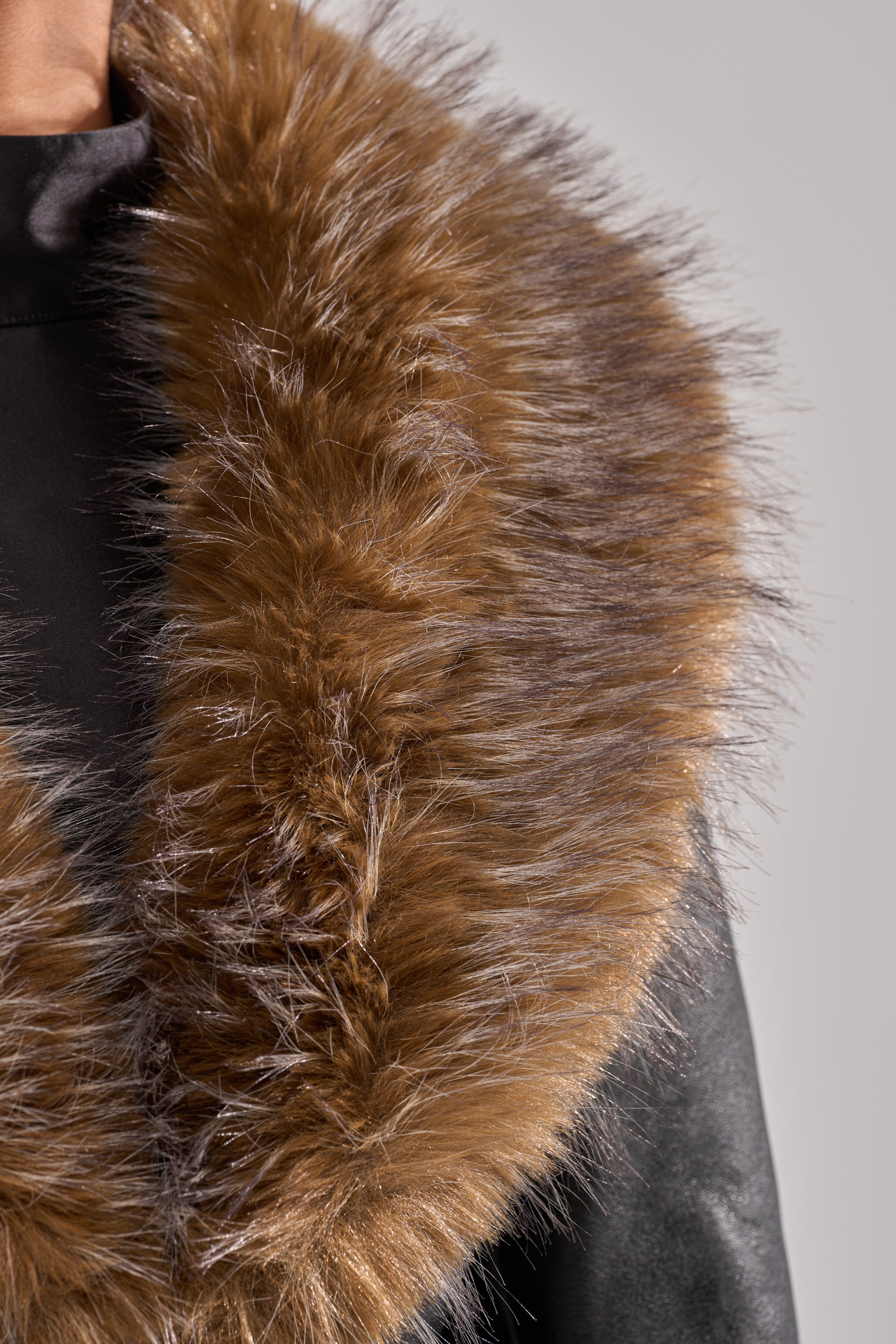 Close-up of a person wearing the VOODOO FUR TRIM TRENCH, featuring a black coat with a large, fluffy brown faux fur trim. The image showcases the texture and details of the trim and upper coat against a light background.