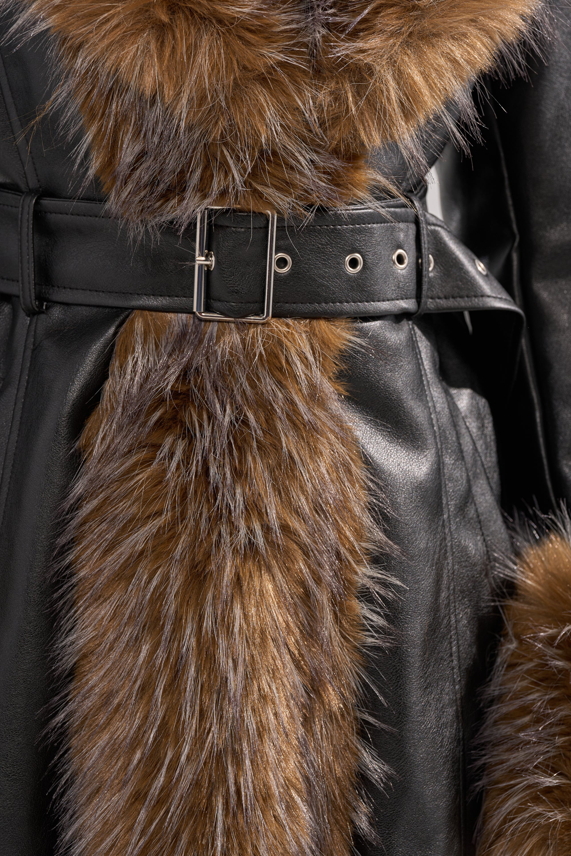 Close-up of the VOODOO FUR TRIM TRENCH: a black faux leather coat with a silver buckle belt and brown fluffy faux fur trim on the collar and front, showcasing the striking contrast between leather and fur.