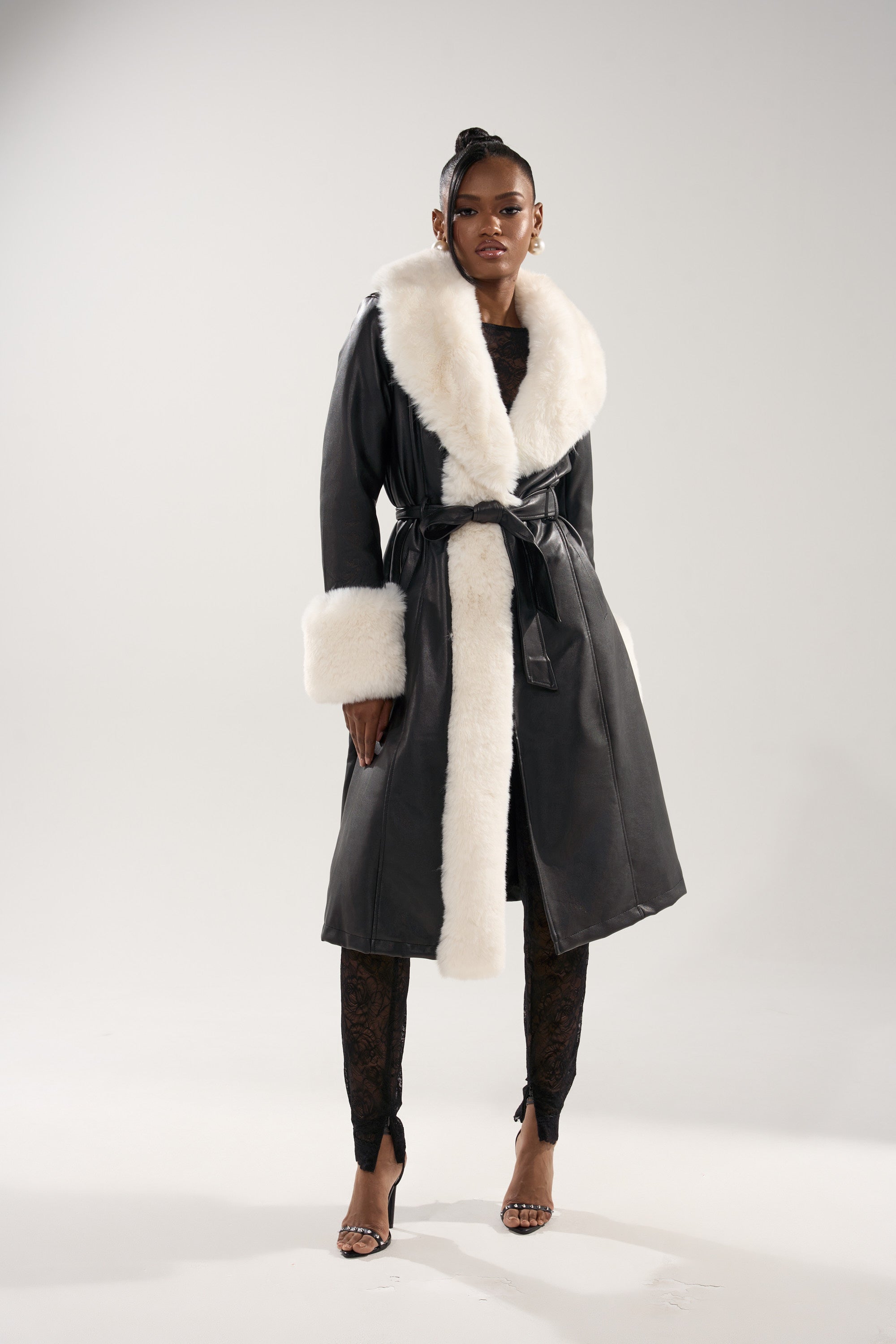A woman poses confidently in the SNOW BUNNY FUR TRIM TRENCH, a black belted coat with white fur collar and cuffs, paired with black lace pants and open-toe heels, set against a plain light background.