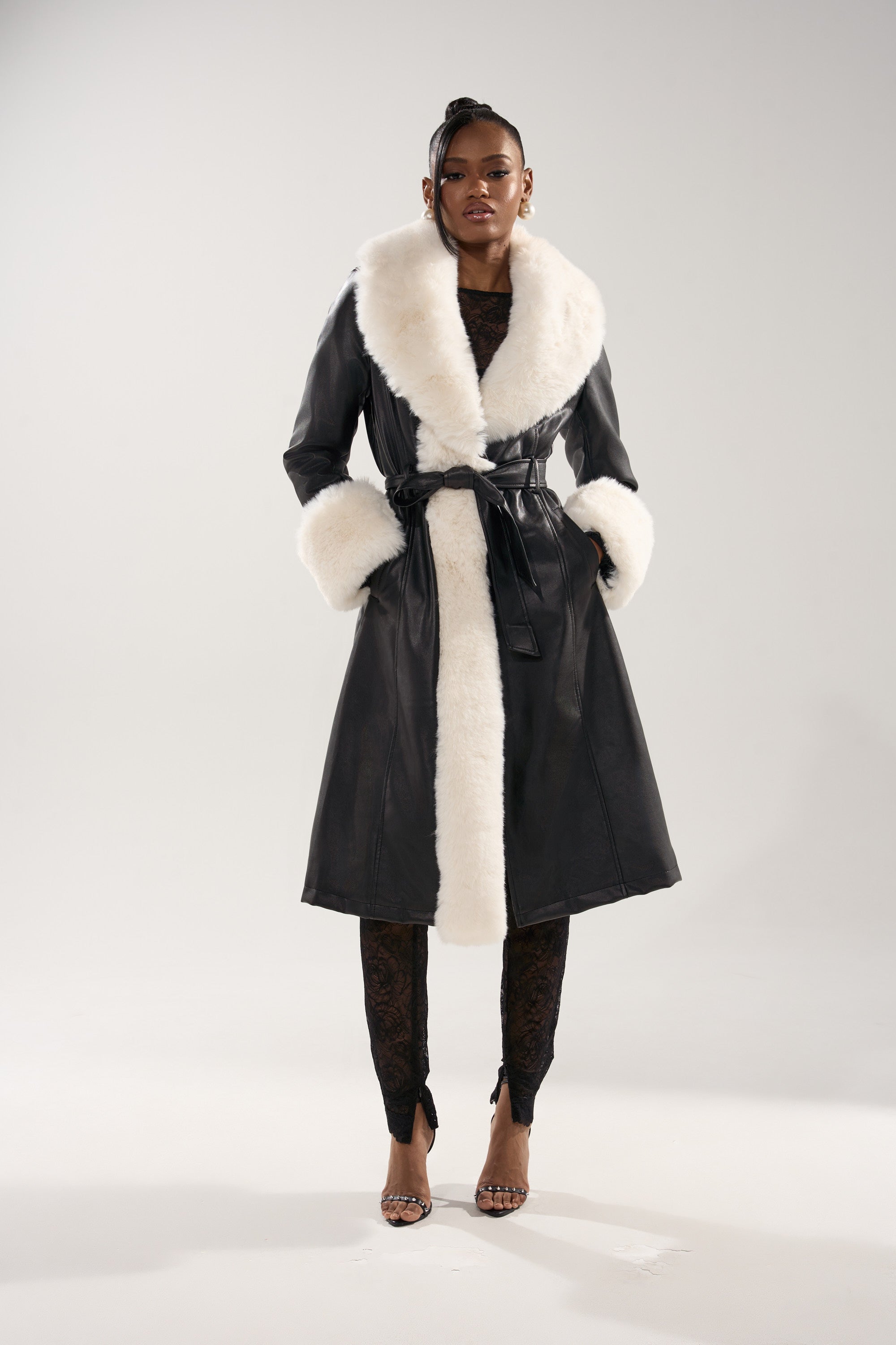 A woman exudes confidence in the SNOW BUNNY FUR TRIM TRENCH, featuring a belted faux leather design with a wide white fur collar and cuffs. She completes the look with black lace pants and open-toed heels against a plain light background.