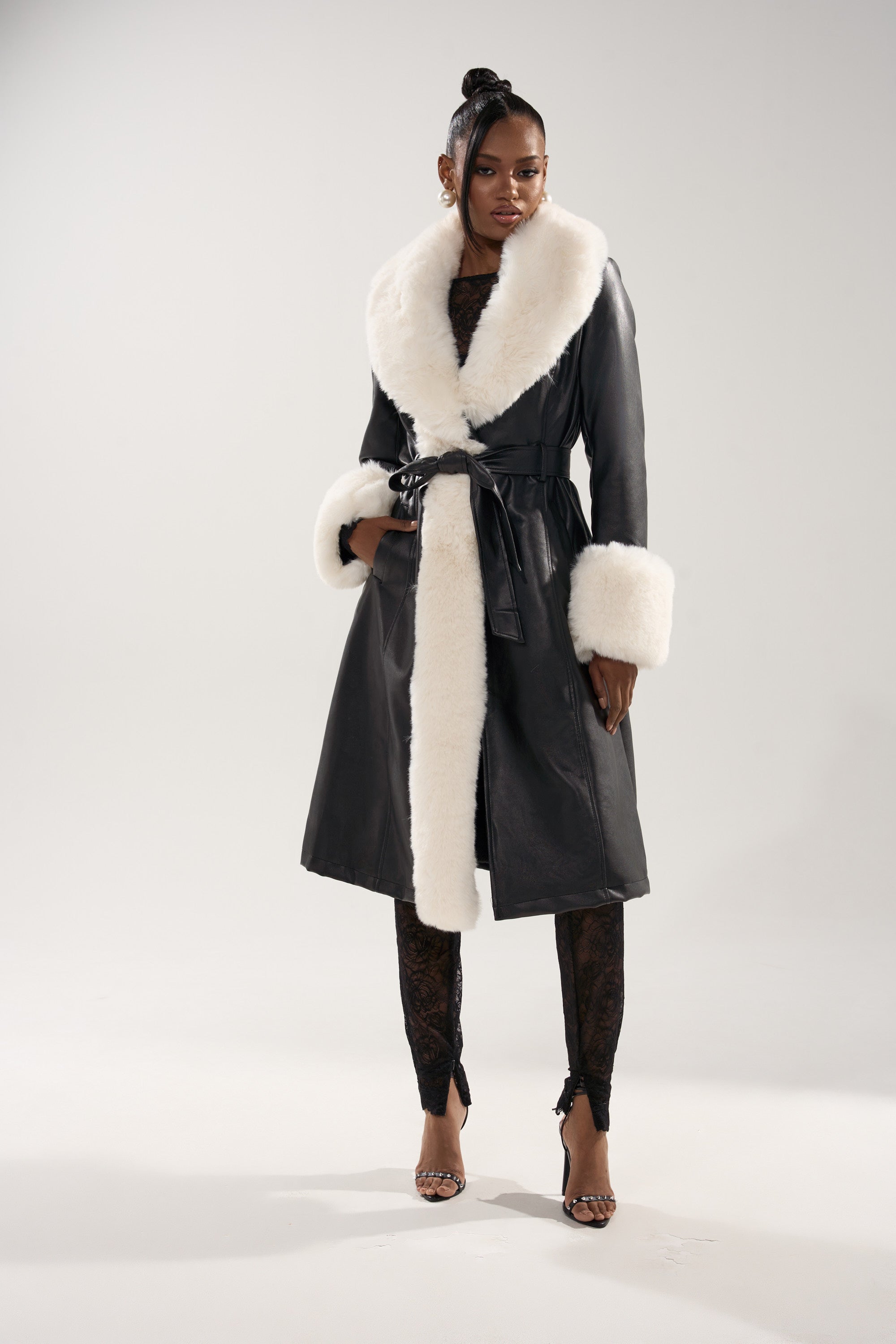 A person wears the SNOW BUNNY FUR TRIM TRENCH, a long black coat with oversized white fur accents on the collar and cuffs, paired with black lace pants and strappy heels, standing against a plain light backdrop.