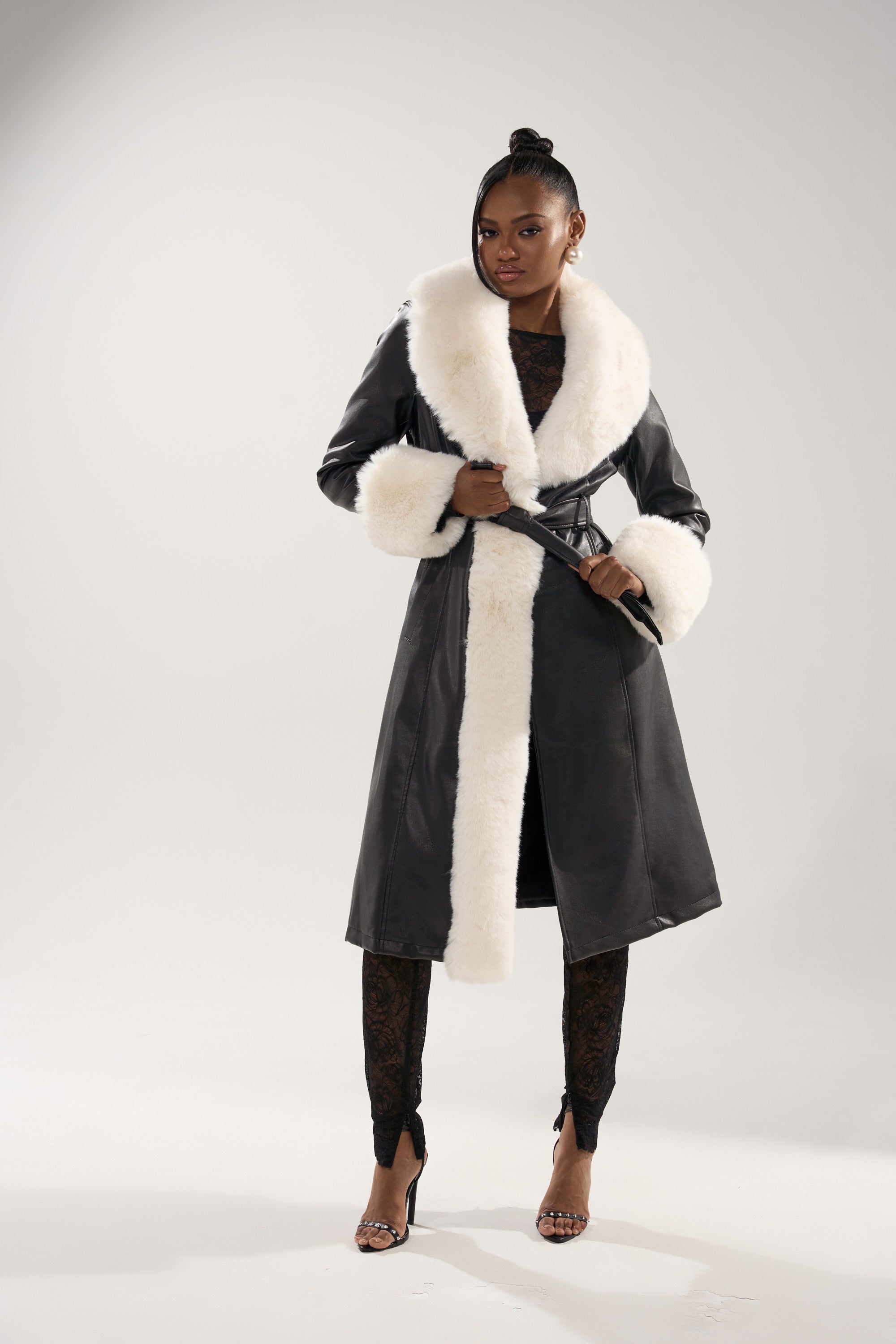 A woman exudes confidence in the SNOW BUNNY FUR TRIM TRENCH, a black faux leather coat with white fur collar, cuffs, and front trim, styled with black lace and strappy heels against a simple light backdrop.