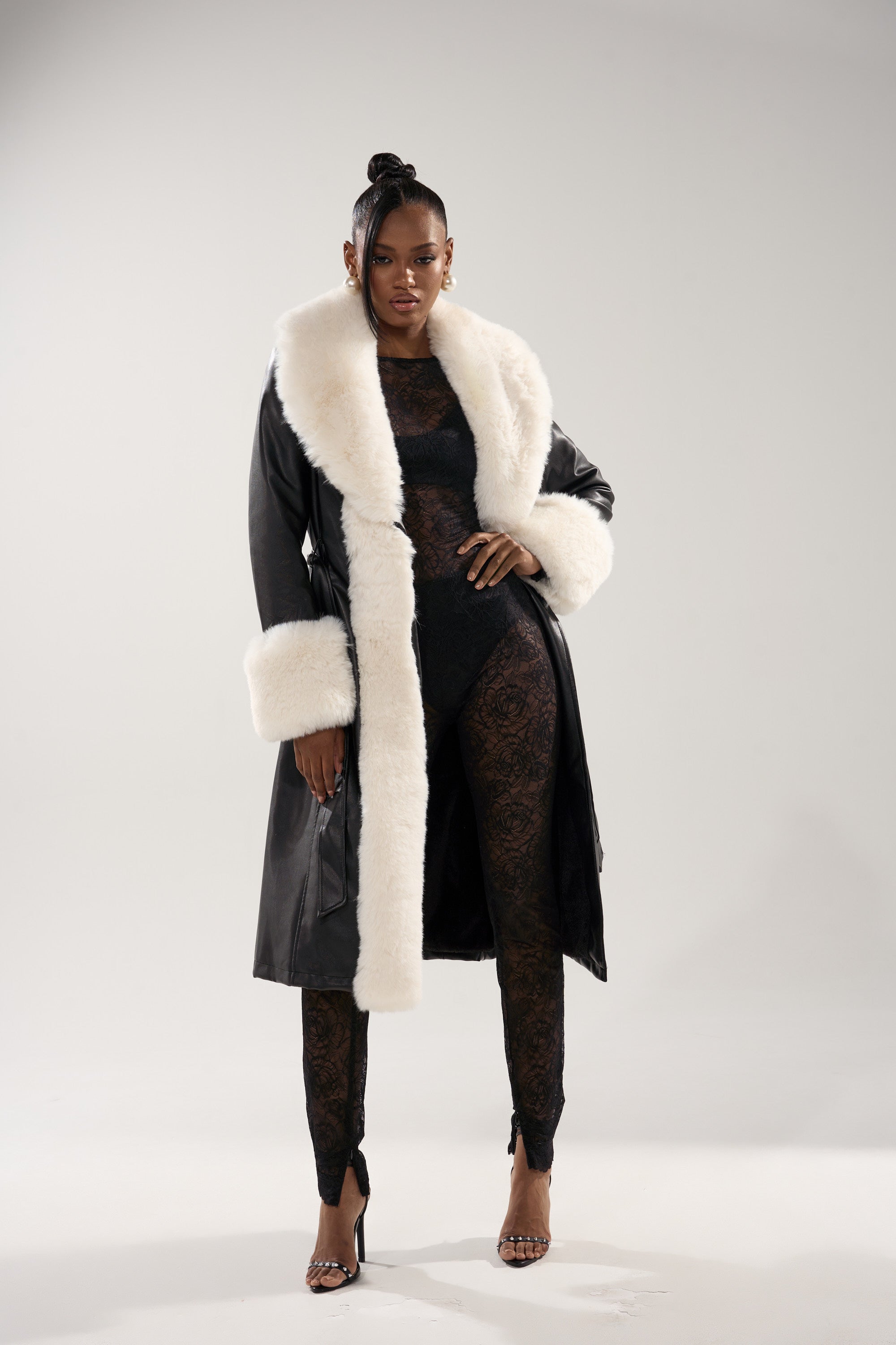 A woman poses confidently in the SNOW BUNNY FUR TRIM TRENCH, a faux leather coat with large white fur trim on the collar and cuffs, layered over a black lace bodysuit and paired with high heels against a plain light background.