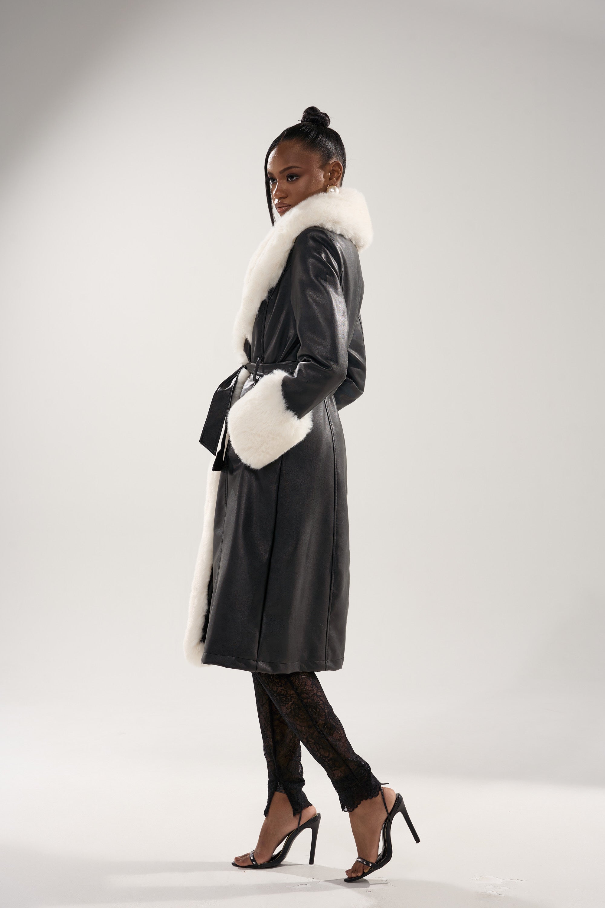 A woman poses confidently in the SNOW BUNNY FUR TRIM TRENCH, a chic faux leather coat with white fur at the collar and cuffs, styled with black lace pants and high-heeled sandals against a plain light background.