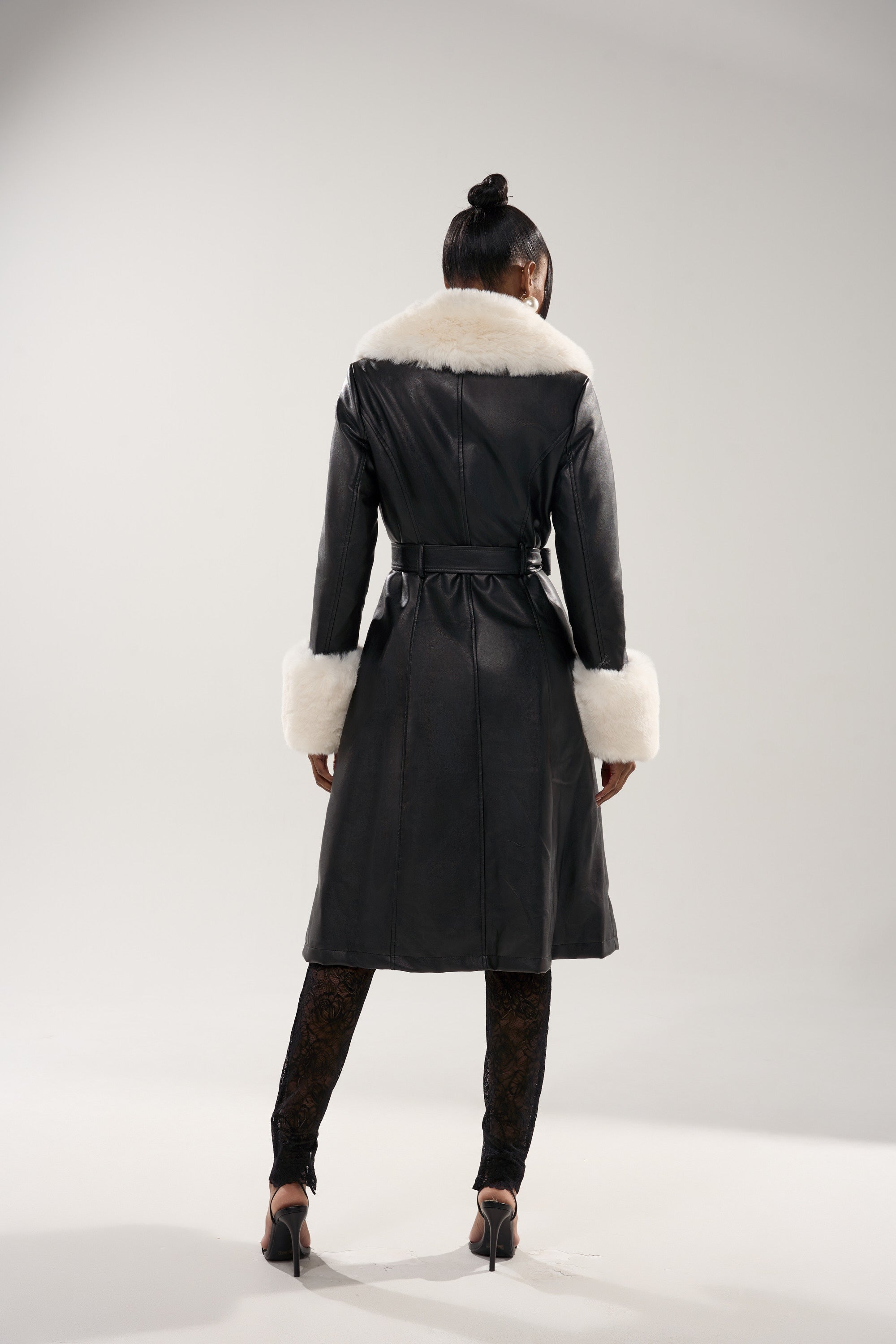 A woman with her hair in a bun models the SNOW BUNNY FUR TRIM TRENCH, featuring a white collar and cuffs. She pairs it with black lace leggings and high heels, standing in a well-lit studio against a plain backdrop.