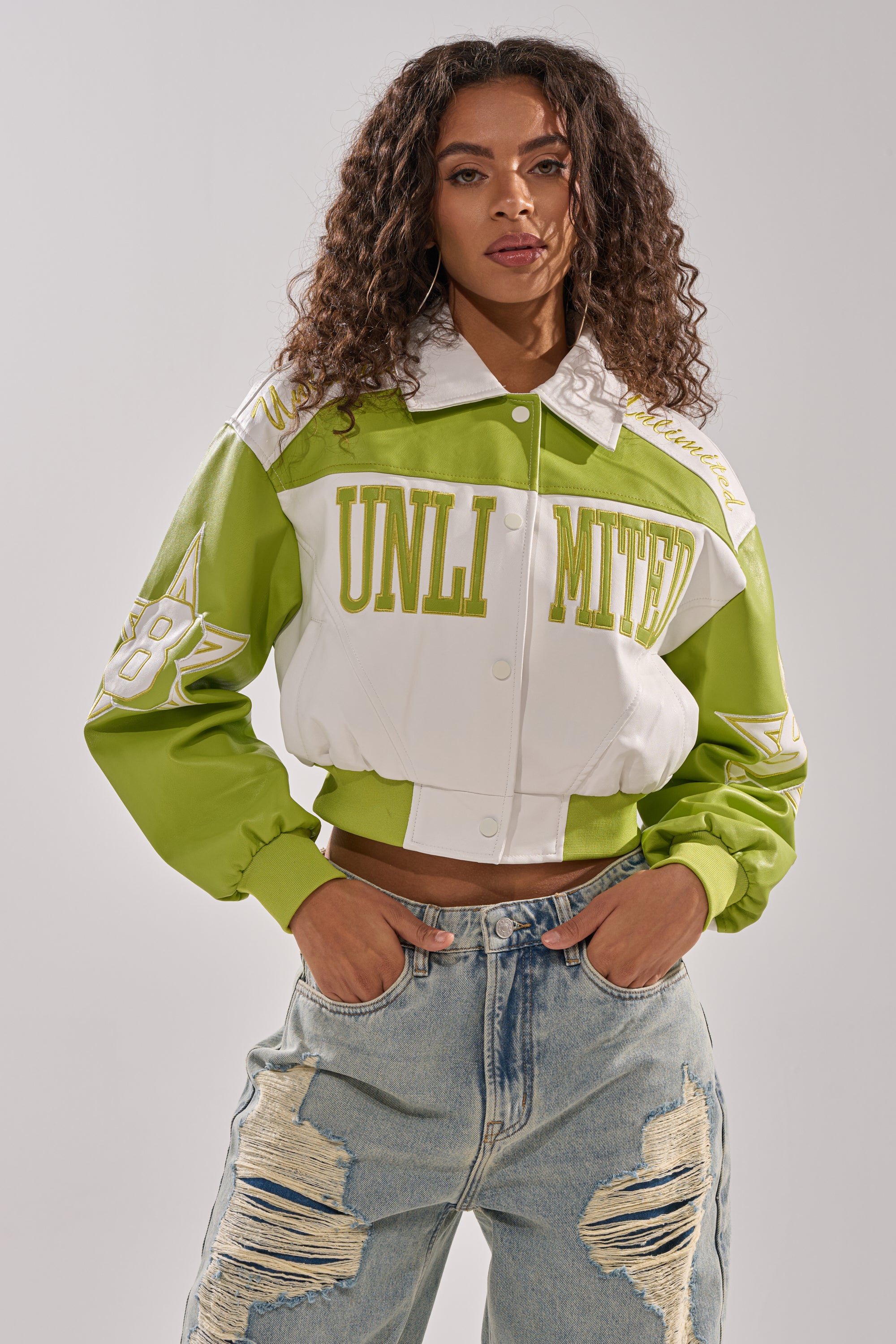 A woman with curly hair rocks the UNLIMITED MOTO BOMBER—a green and white varsity-style jacket with "UNLIMITED" on the front—paired with ripped, light-wash jeans, exuding effortless confidence with her hands in her pockets.