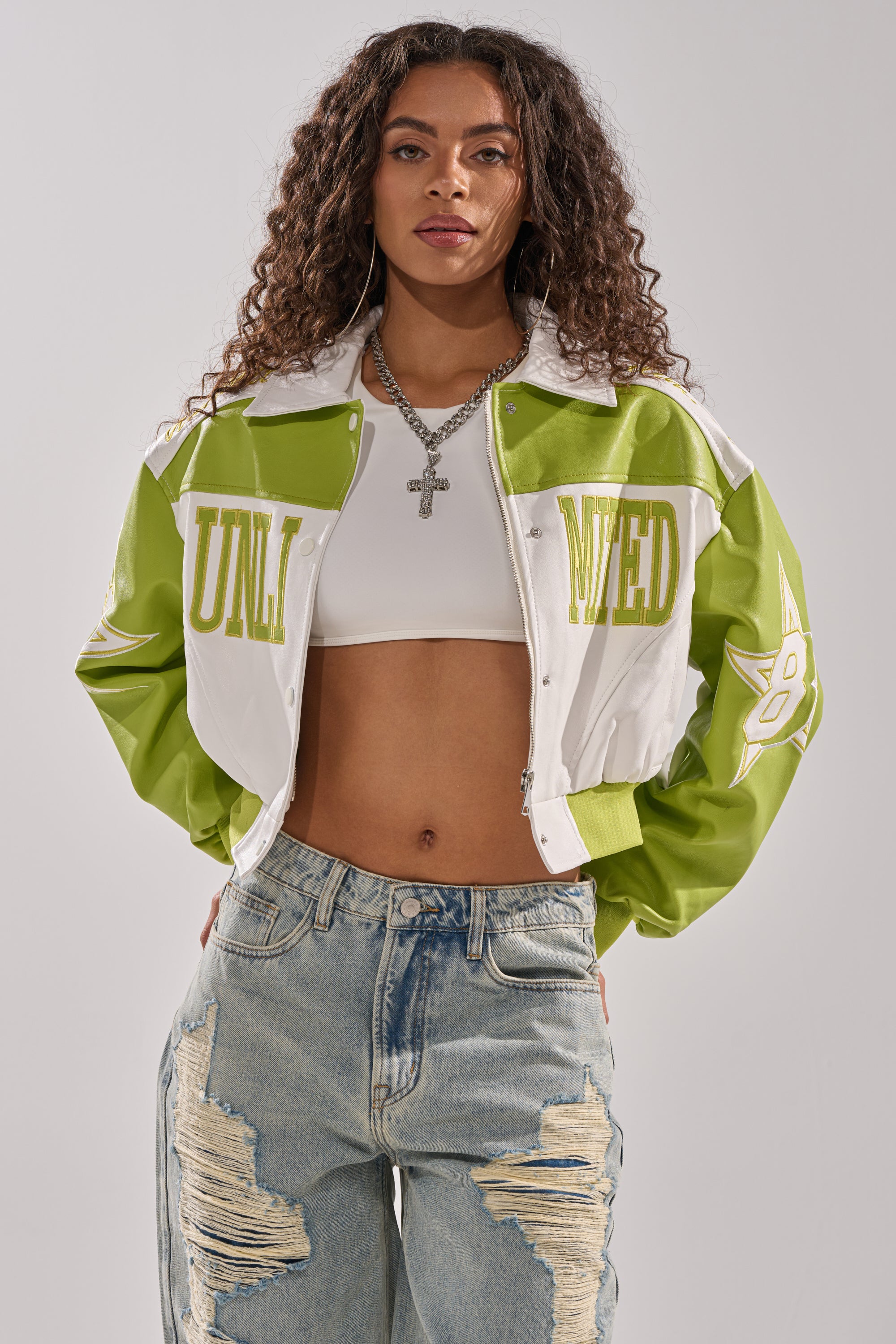A woman with long curly hair wears the UNLIMITED MOTO BOMBER, a green and white cropped varsity jacket, over a white crop top with layered necklaces and ripped blue jeans. She stands confidently against a plain background.
