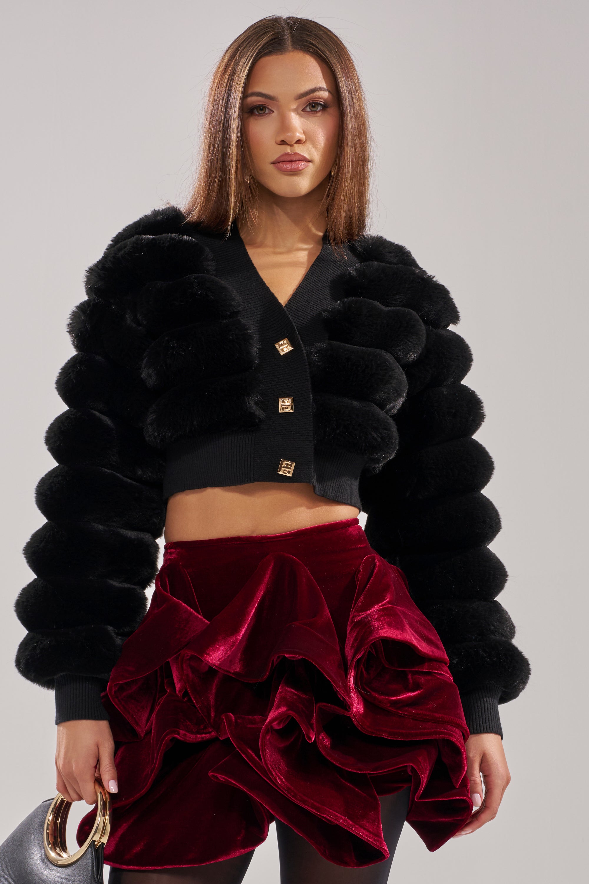 A woman wears the WILD HEART FAUX FUR CARDIGAN IN BLACK with a deep red ruffled velvet mini skirt, holding a black handbag with a gold handle. She has straight, shoulder-length hair and a neutral expression. 0