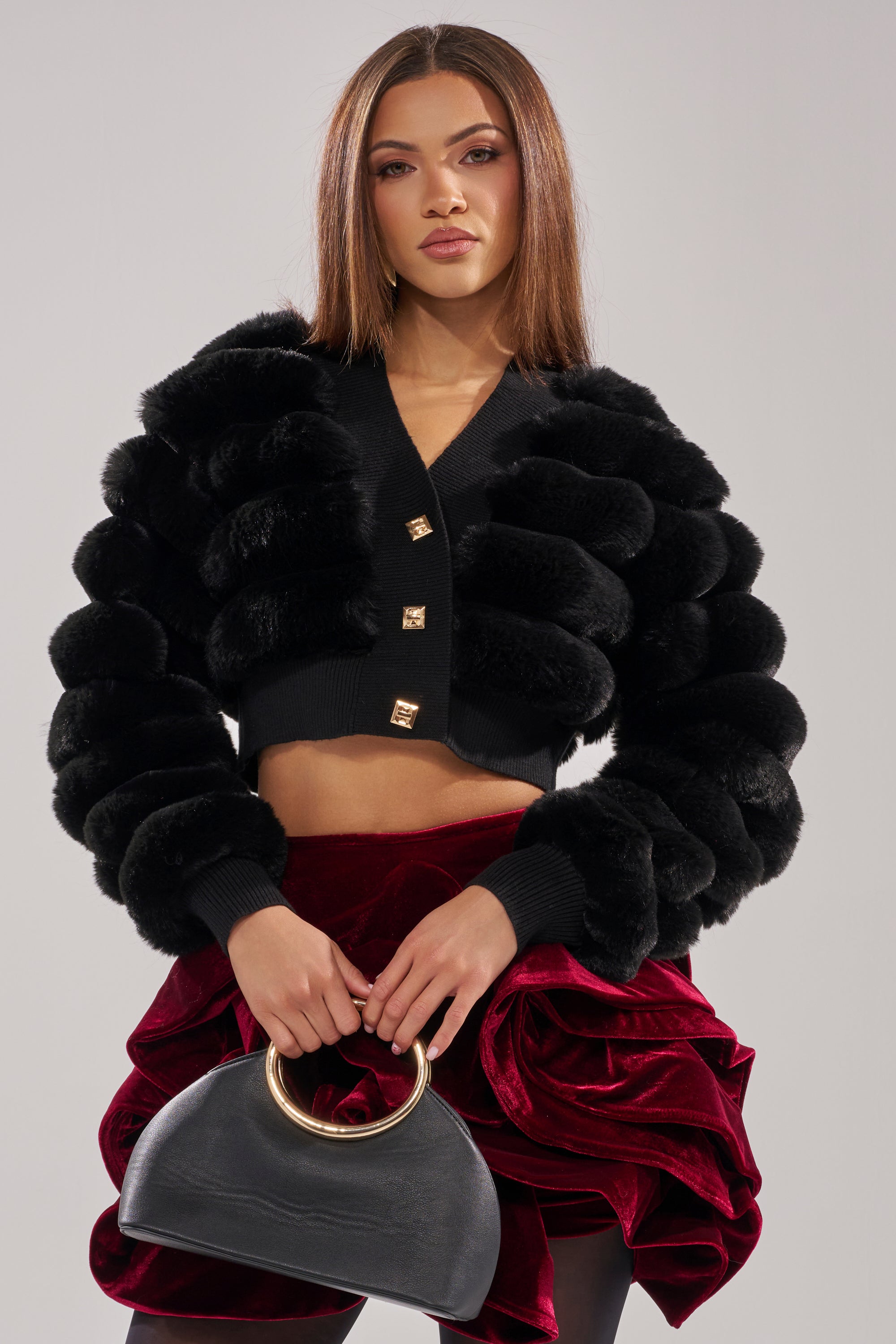 A woman poses confidently in the WILD HEART FAUX FUR CARDIGAN IN BLACK, paired with a red velvet ruffled mini skirt and a black gold-handle handbag. She has straight brown hair and natural makeup. 2