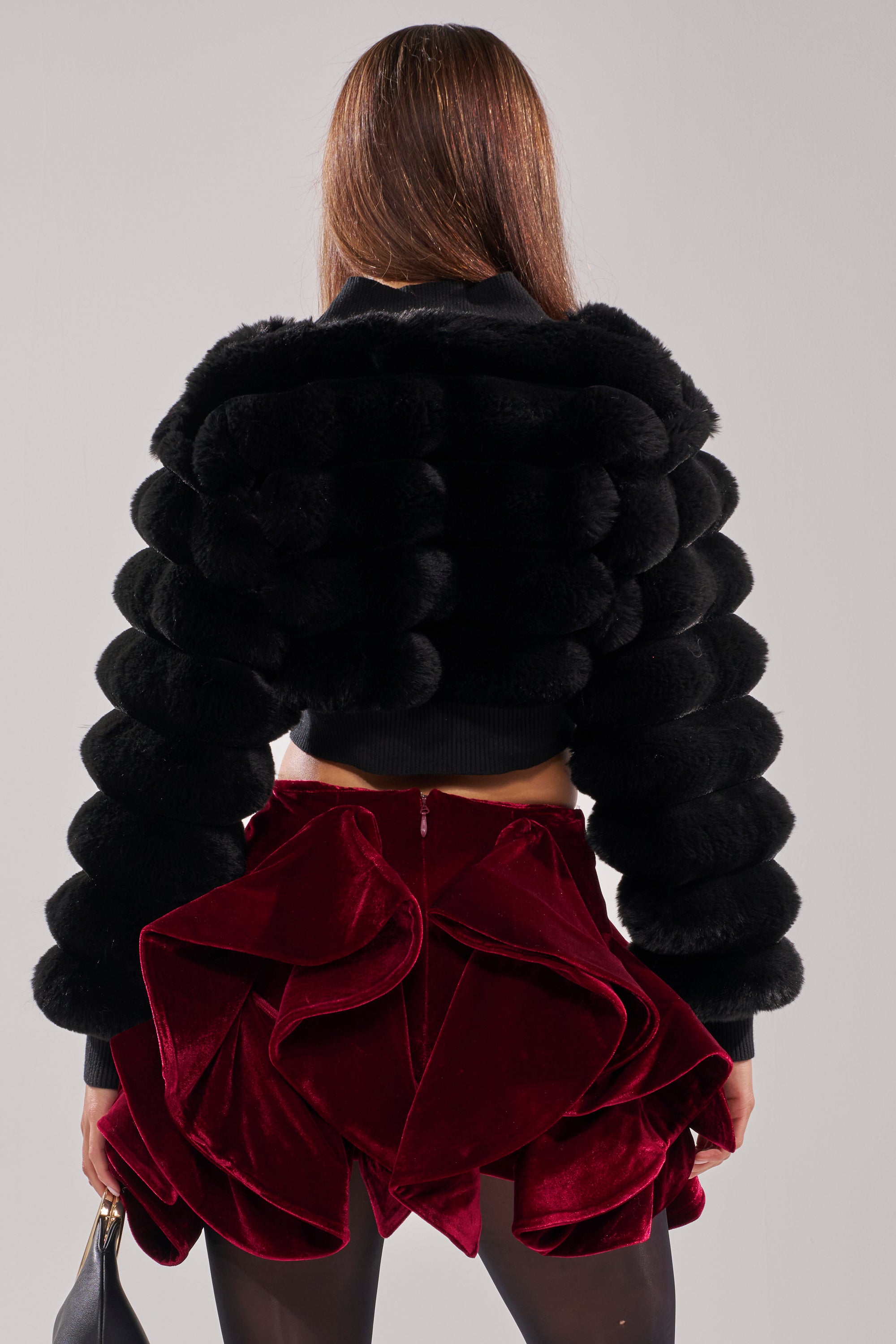 A person with straight brown hair models the WILD HEART FAUX FUR CARDIGAN IN BLACK, styled with a short ruffled deep red velvet skirt, photographed from behind against a plain light background. 4