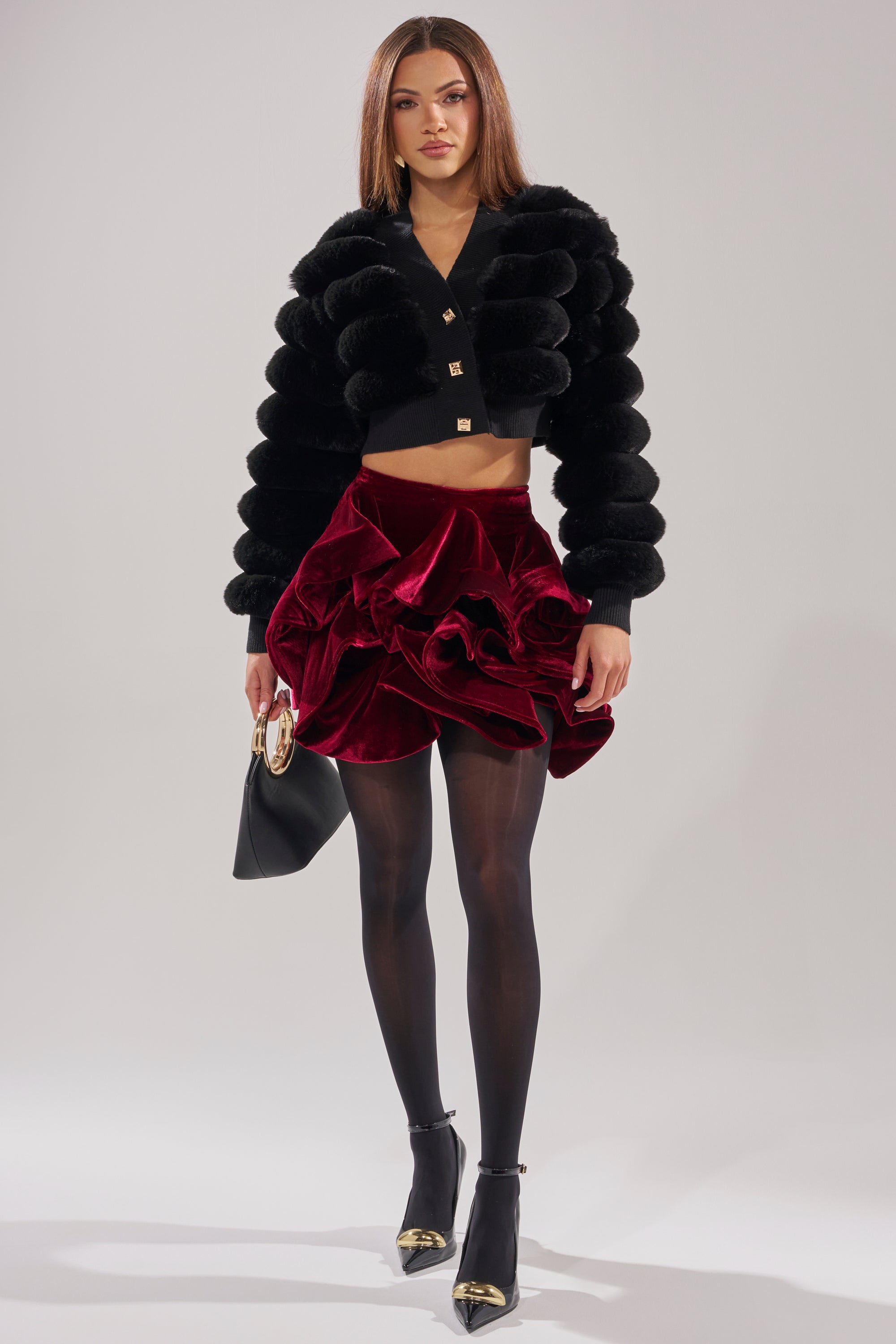 A woman poses in the WILD HEART FAUX FUR CARDIGAN IN BLACK, paired with a red velvet ruffled mini skirt, sheer black tights, black heels with gold tips, and holding a black handbag against a simple light background. 5