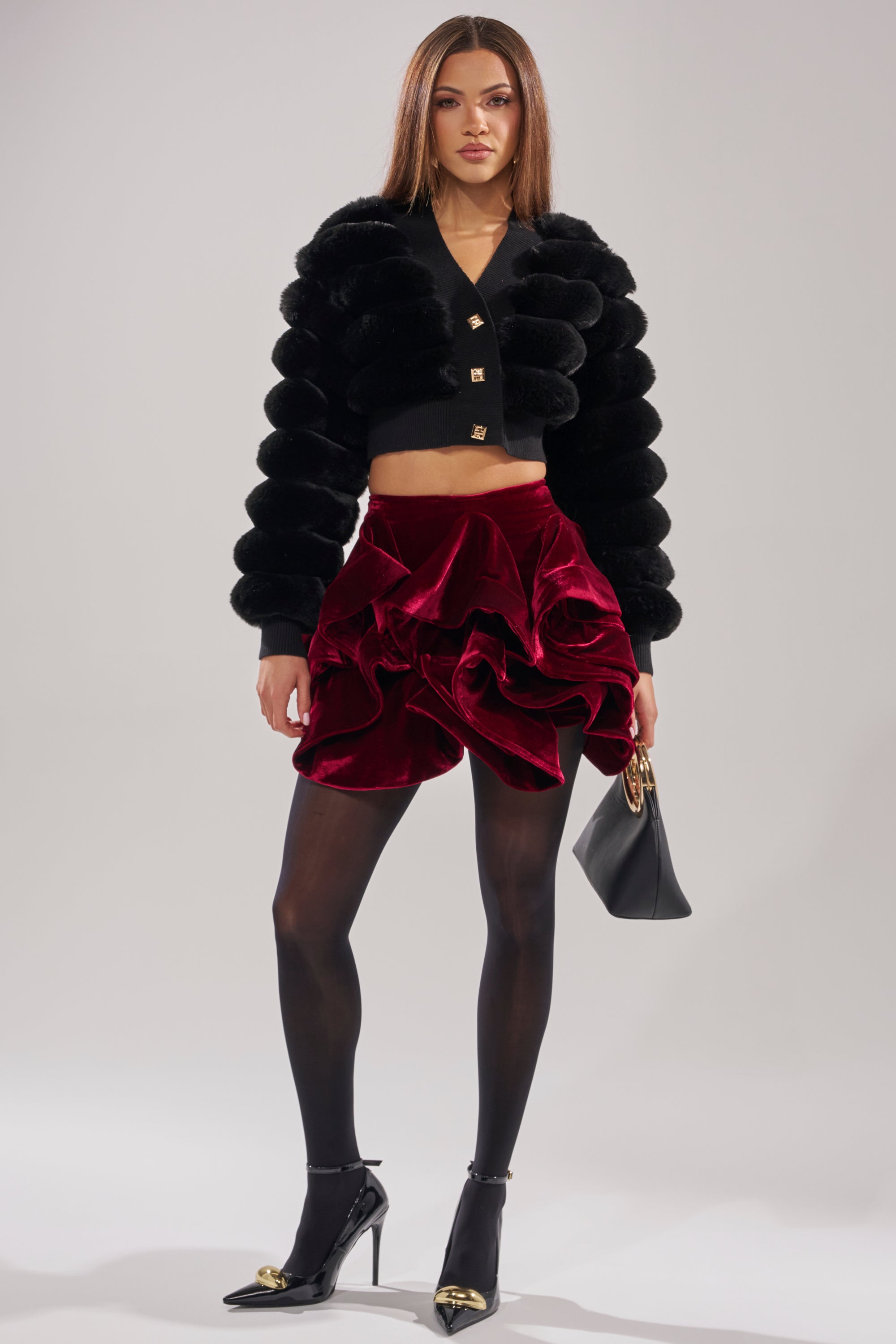 A woman poses in the WILD HEART FAUX FUR CARDIGAN IN BLACK, styled with a ruffled red velvet mini skirt, black sheer tights, gold-detailed black heels, and a small black handbag. 6