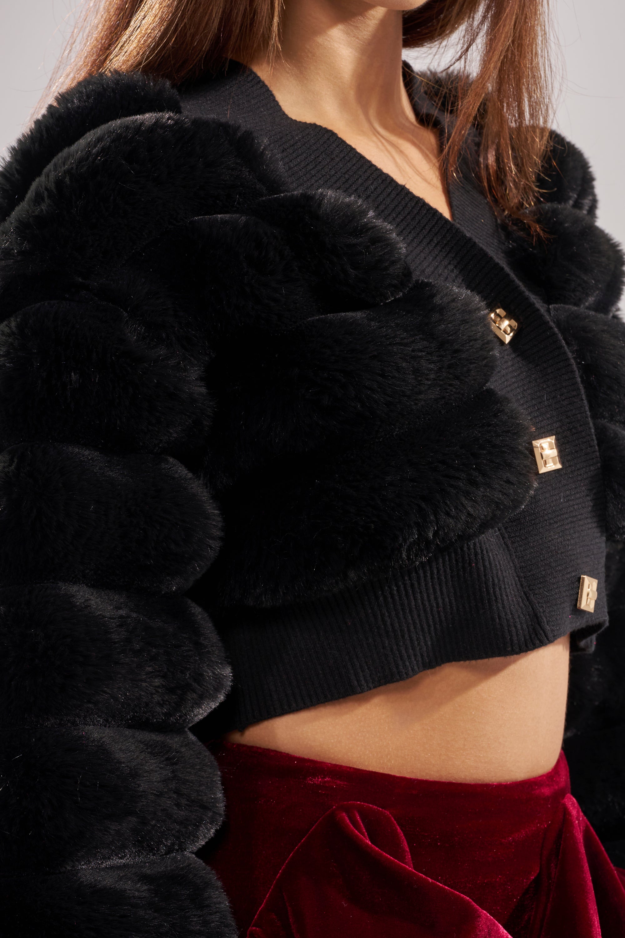 A woman wears the WILD HEART FAUX FUR CARDIGAN IN BLACK, featuring gold square buttons, paired with a deep red velvet skirt with a large bow. The chic AKIRA Label look is shown in a photo cropped from shoulders to waist. 8