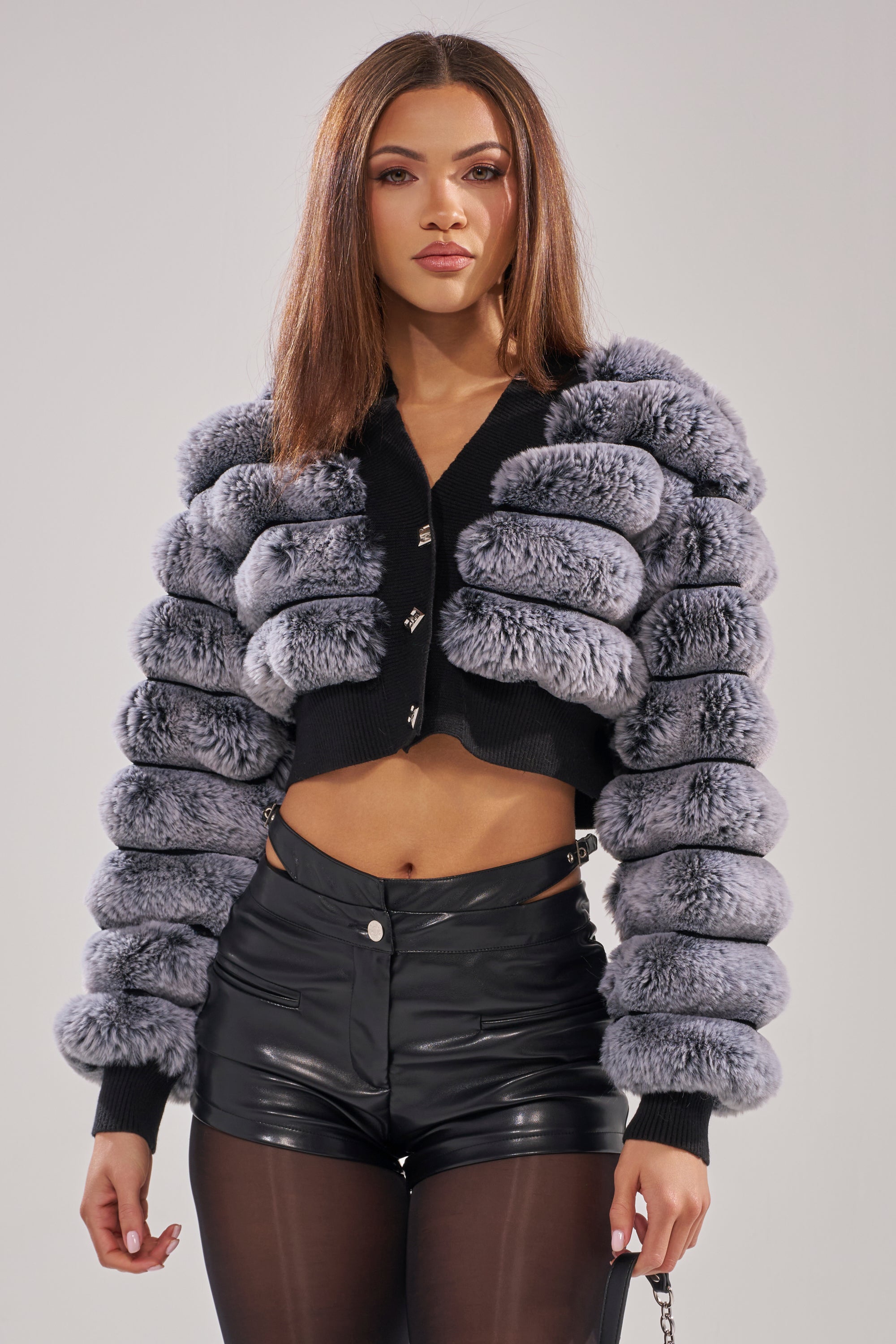 A woman with straight brown hair exudes modern glamour in the WILD HEART FAUX FUR CARDIGAN, cropped and puffy in gray, layered over a black top, high-waisted black leather shorts, and sheer black tights against a plain light background.