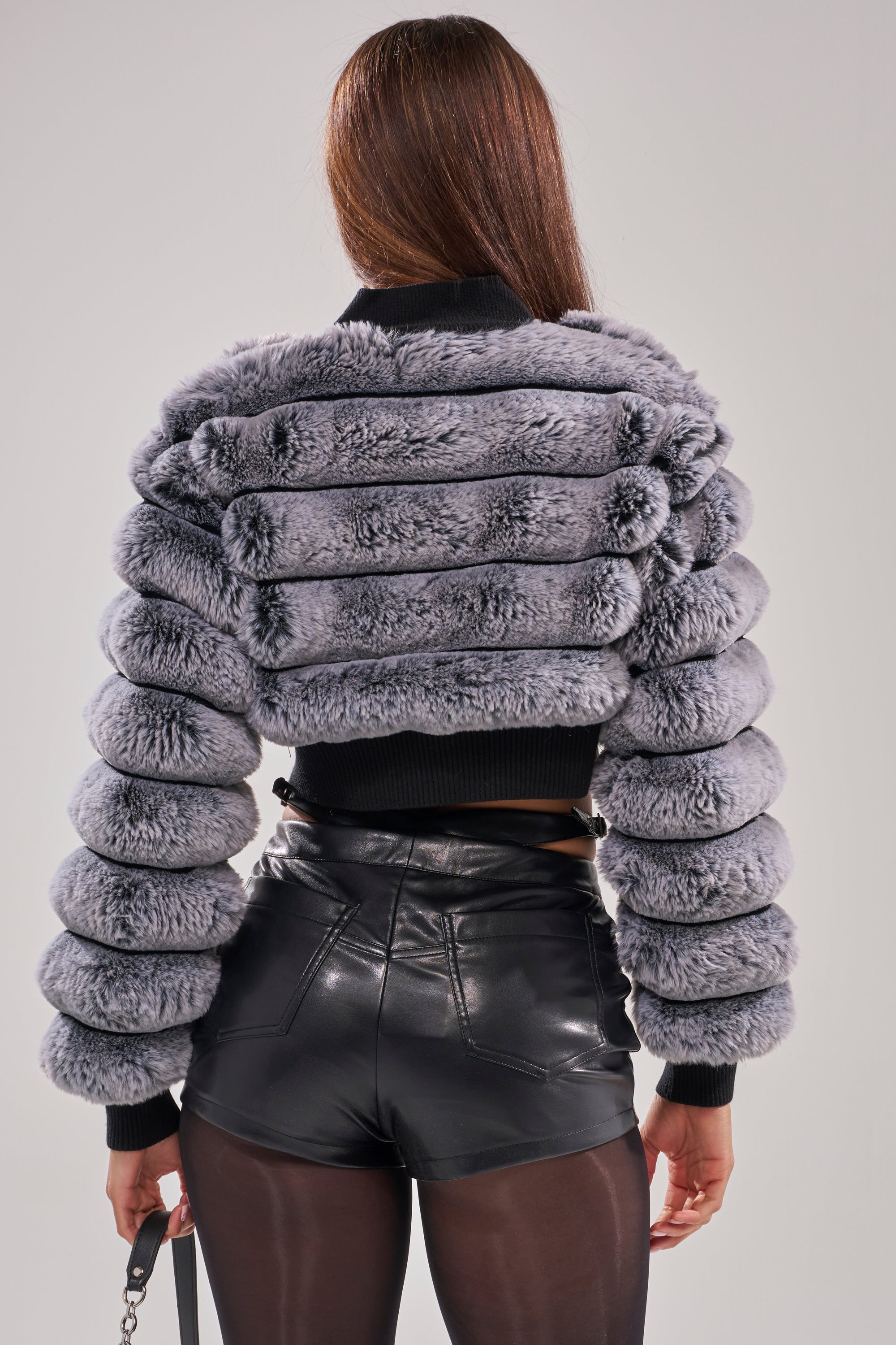 A person with long brown hair is shown from the back against a plain background wearing the AKIRA Label WILD HEART FAUX FUR CARDIGAN, black high-waisted leather shorts, and sheer black tights.